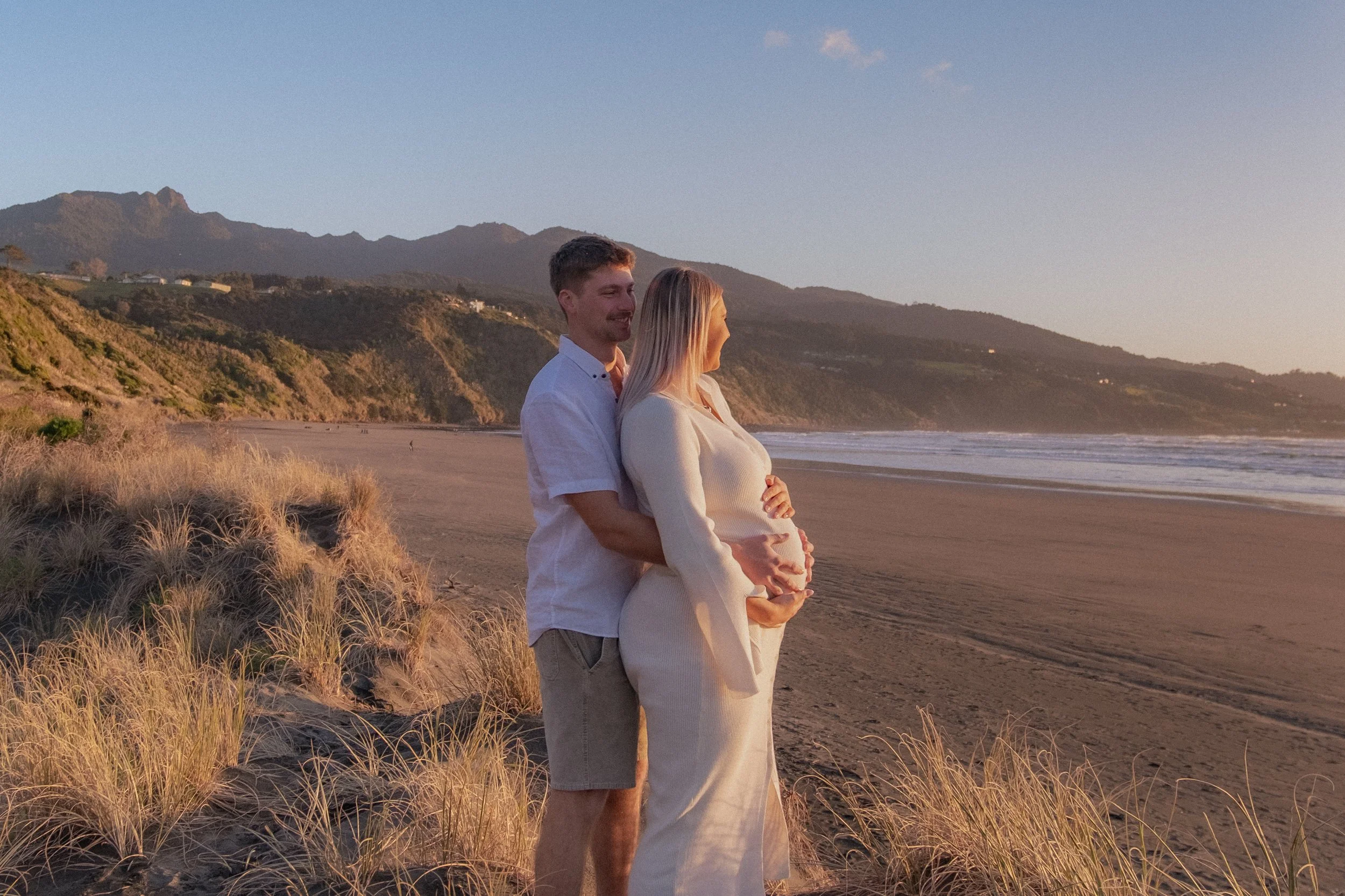 A couple on the beach at sunset, with the man standing behind the pregnant woman, both smiling and looking at each other, surrounded by sand dunes and coastal landscape. Maternity Shoot. Raglan New Zealand