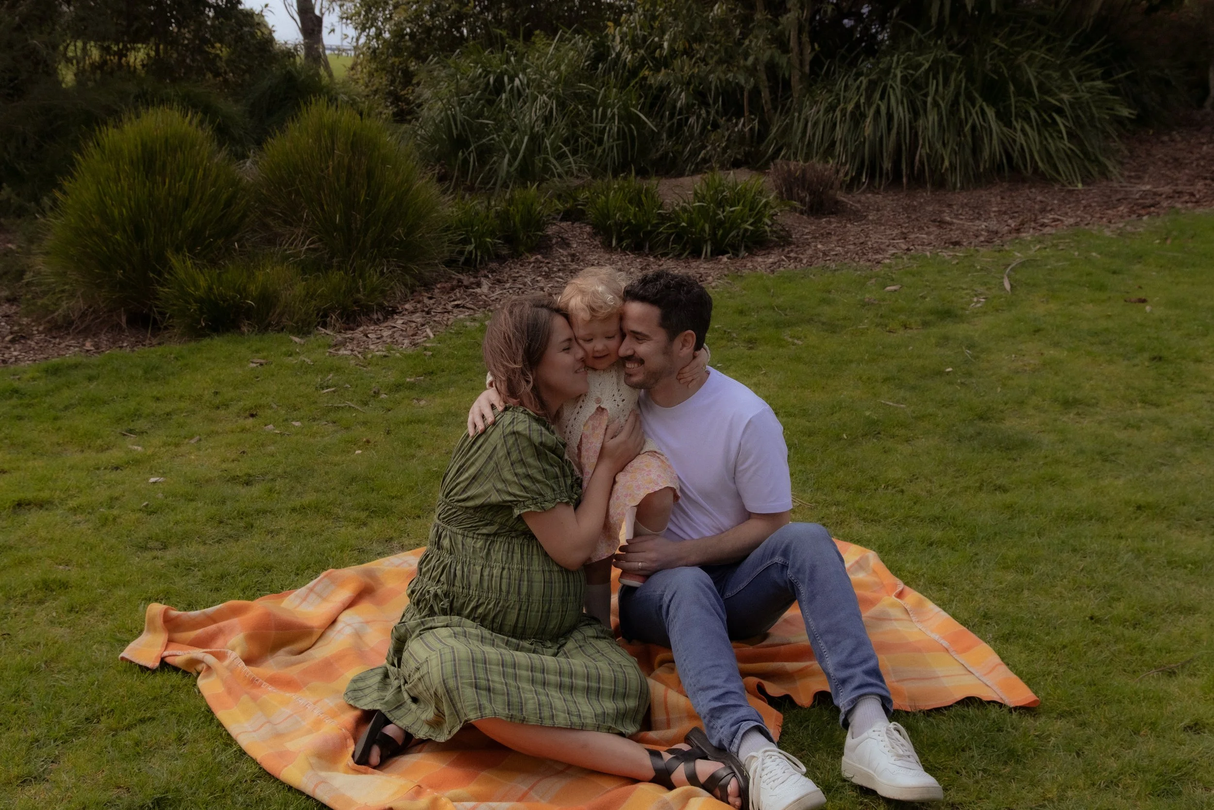 Family of three sitting on an orange checkered blanket in a park, smiling and hugging each other. Family photographer, Maternity photography, Hamilton New Zealand Photographer