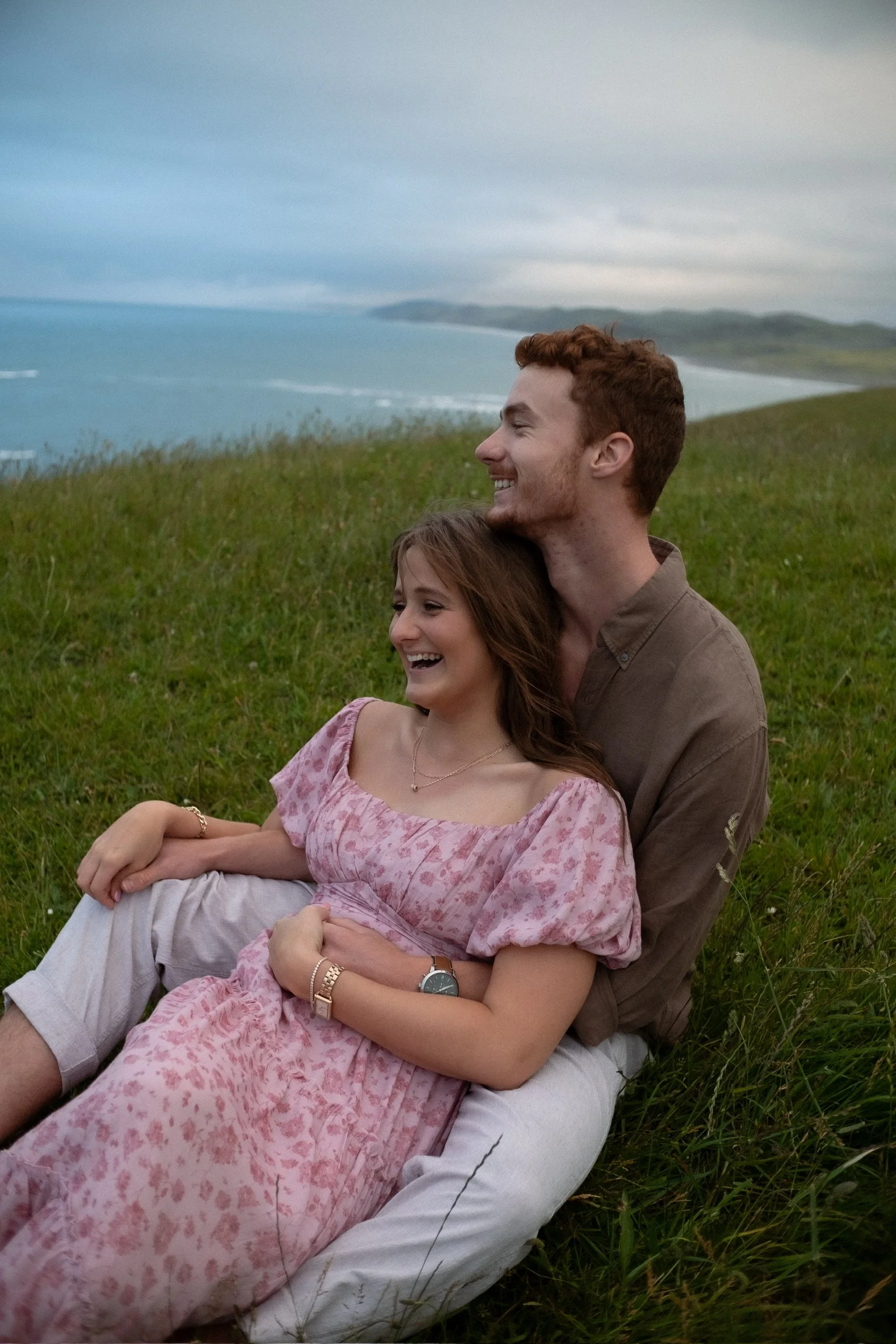 A young couple sitting on grass outdoors, smiling and laughing, with a scenic view of the ocean and hills in the background. Couples photography Raglan New Zealand. Engagement photography Hamilton New Zealand. Whangarei New Zealand Couples photograph