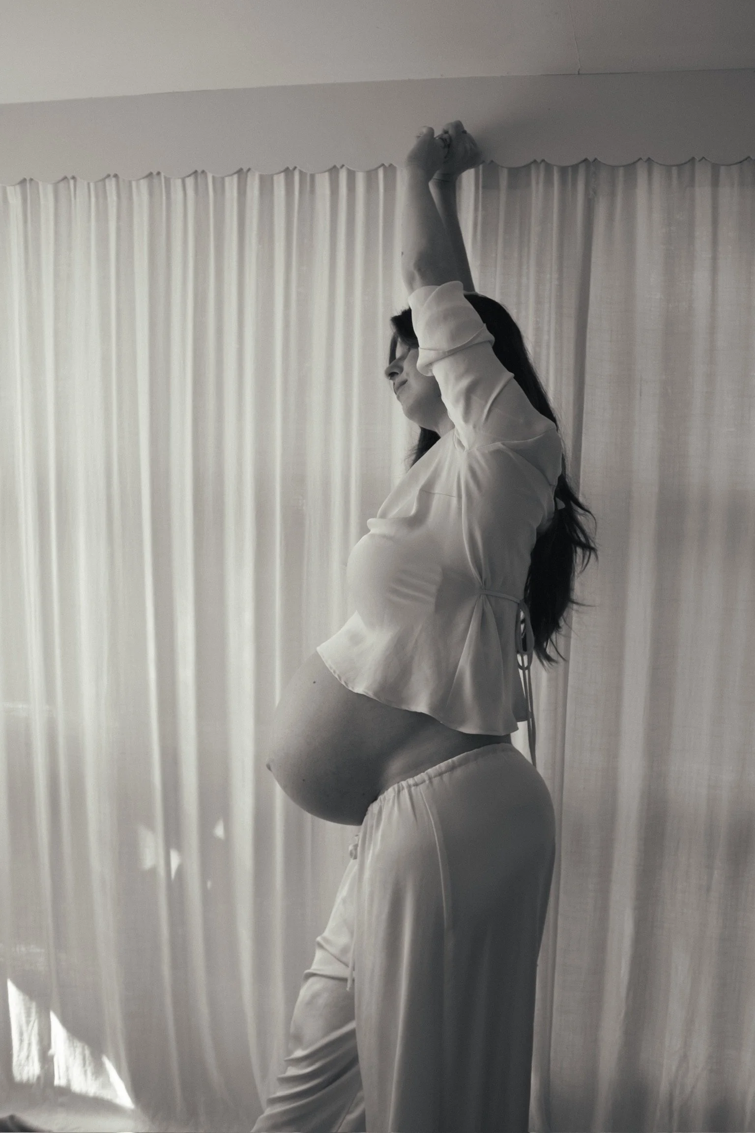 A pregnant woman with long dark hair, standing sideways, with one arm raised above her head, in front of a curtain backdrop. Maternity Photography. Hamilton New Zealand Photographer