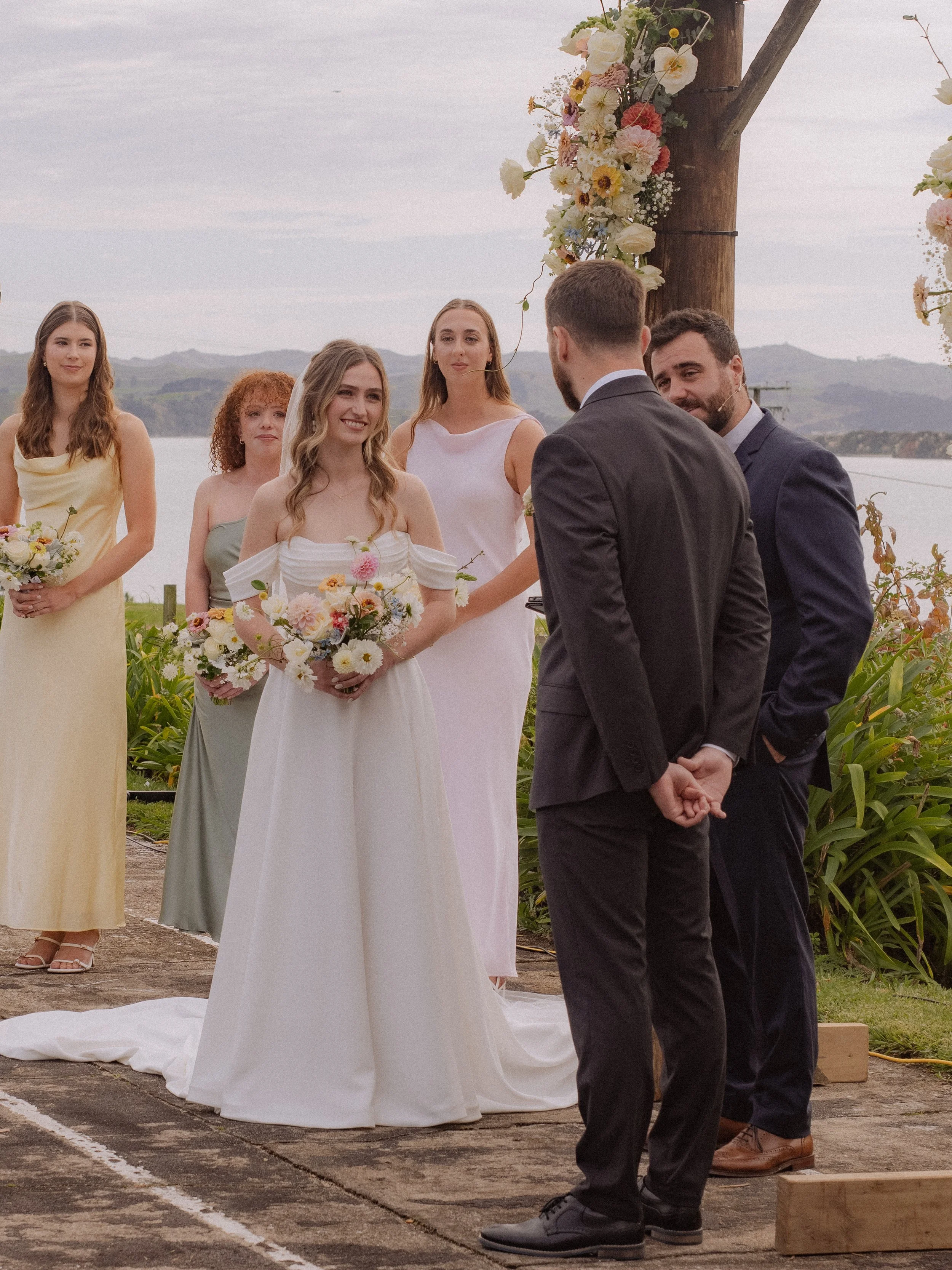 A wedding ceremony outdoors by a lake, with the bride in a white dress holding a bouquet and the groom in a black suit, standing before friends or bridesmaids and groomsmen. The backdrop includes a floral arch and natural scenery with mountains and w