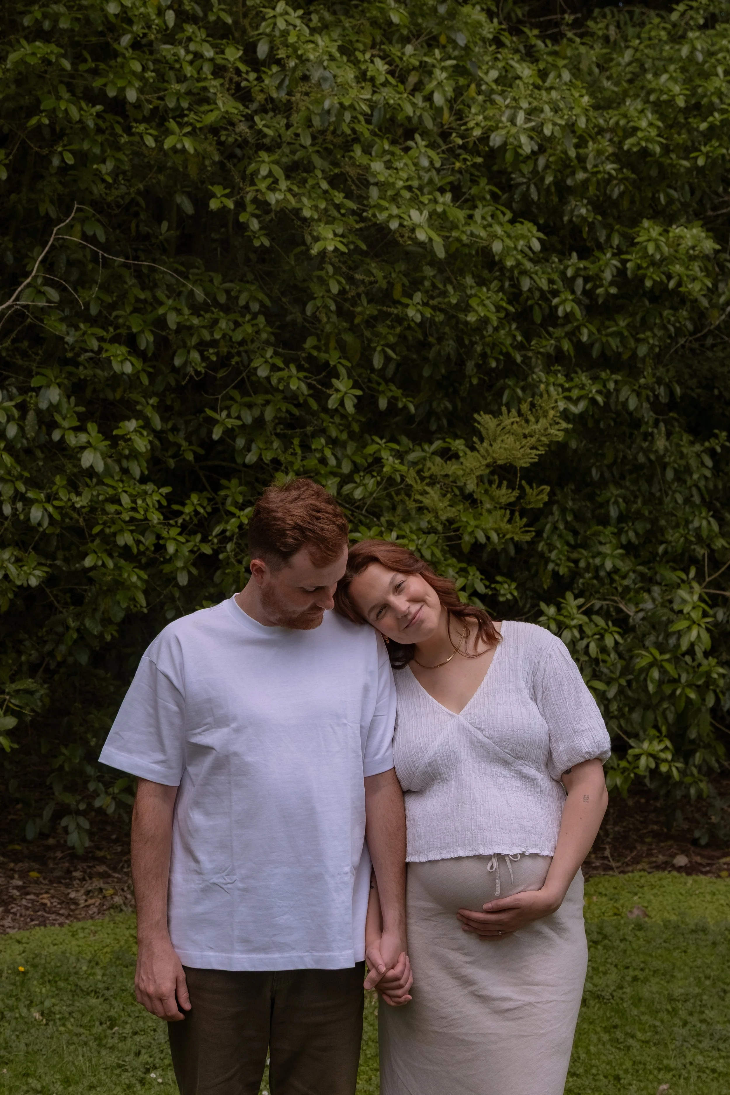 A man and a pregnant woman holding hands, with the woman resting her head on the man's shoulder in a natural outdoor setting. Maternity Photography, Hamilton New Zealand Photographer