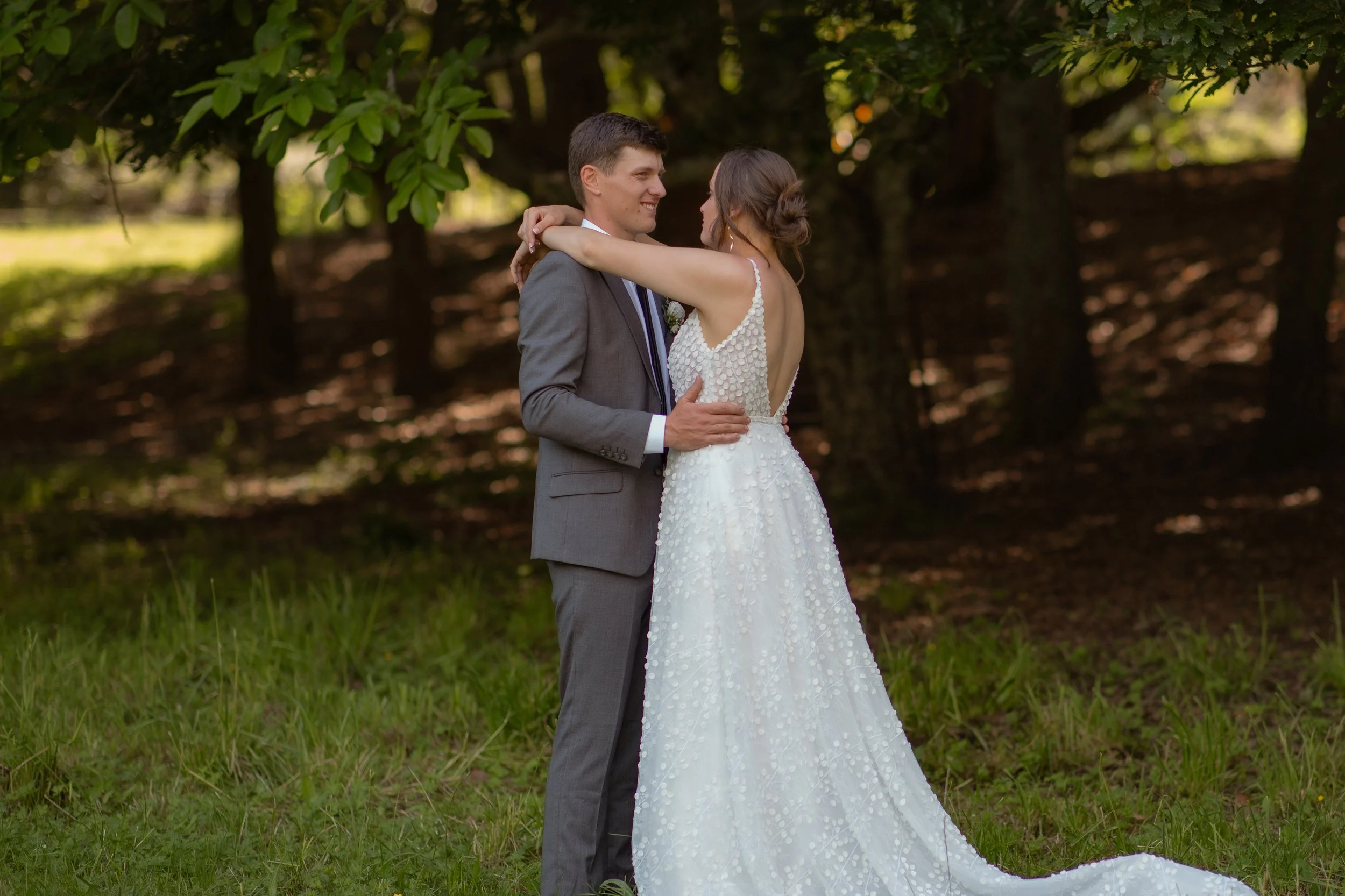 A bride and groom embrace during their wedding in a wooded outdoor setting, with the bride in a white gown and the groom in a gray suit.  Wedding Photography Waikato New Zealand. Wedding Photography Hamilton New Zealand. Northland Wedding Photography