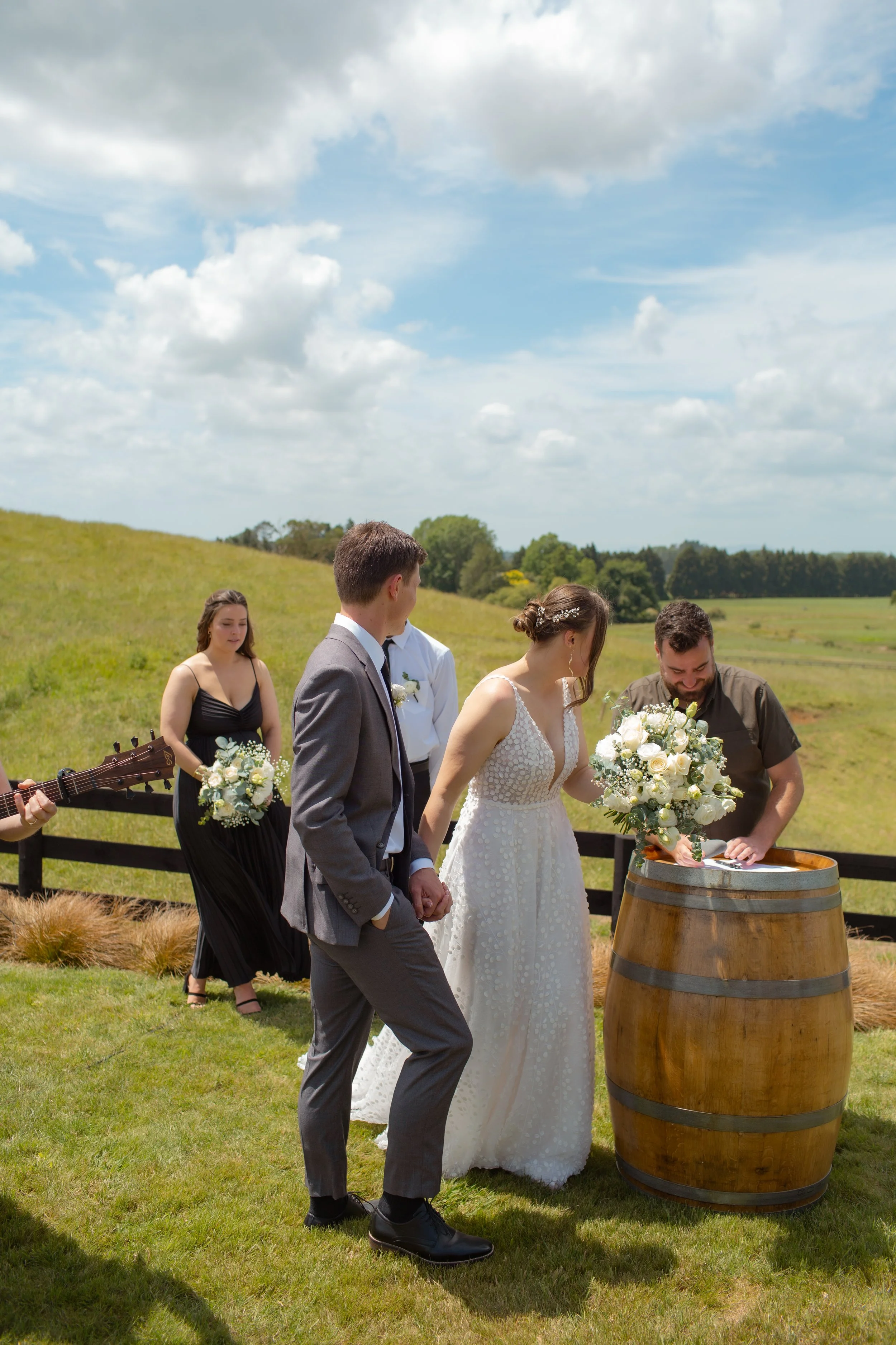 A wedding ceremony outdoors with a bride, groom, officiant, a bridesmaid, and a musician, set amid green fields and blue sky with clouds.  Wedding Photography Waikato New Zealand. Wedding Photography Hamilton New Zealand. Northland Wedding Photograph