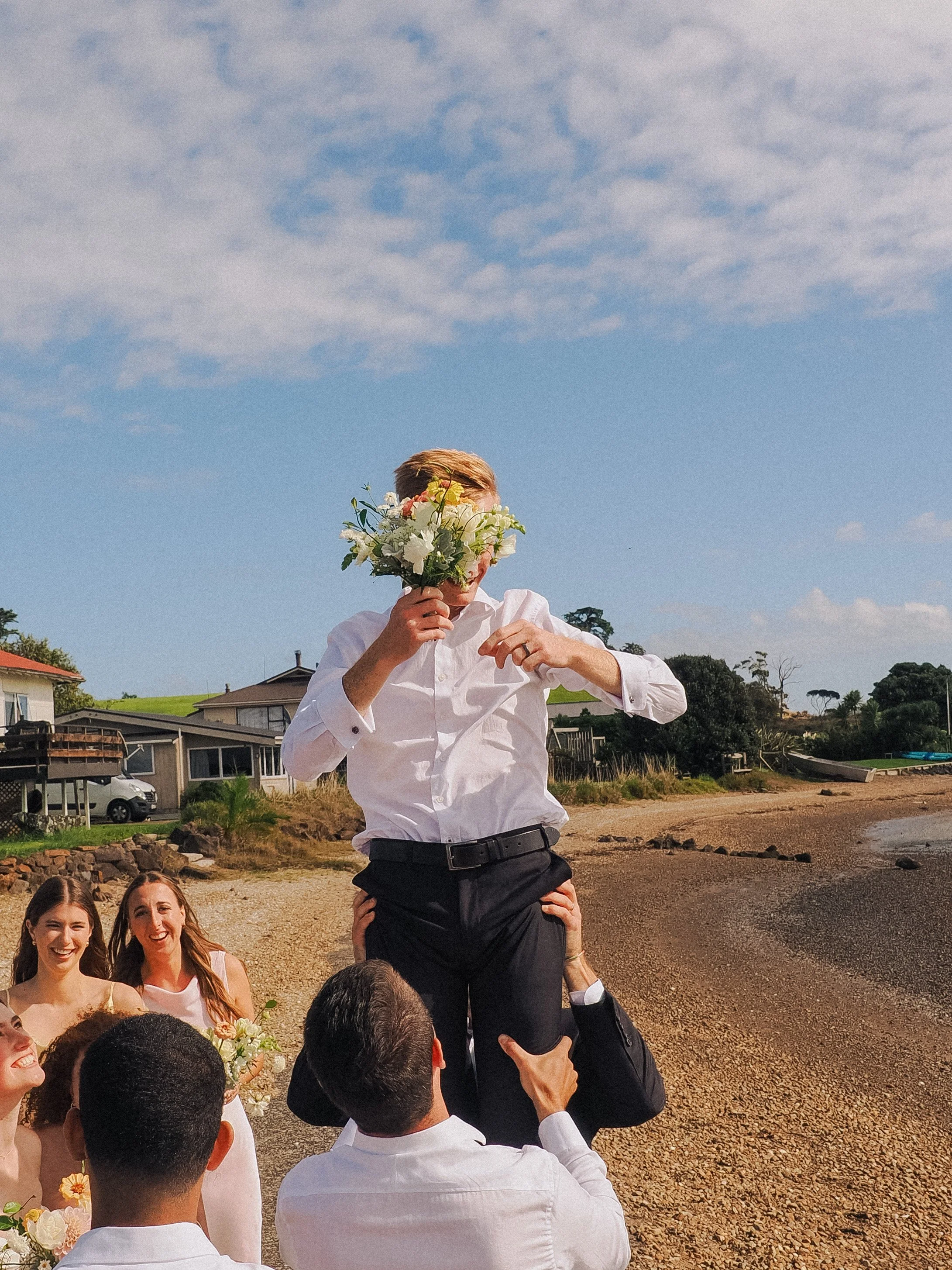 A man holding a bouquet of flowers over his face while being lifted up by another person. Two women and another man are smiling in the background at an outdoor event near the beach with houses and trees in the distance.