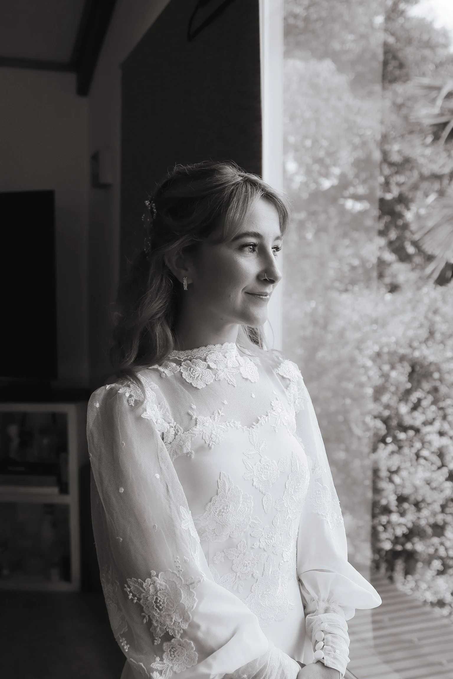 A young woman in a wedding dress looking out a window, smiling softly, in black and white. Wedding Photography Waikato New Zealand. Wedding Photography Hamilton New Zealand