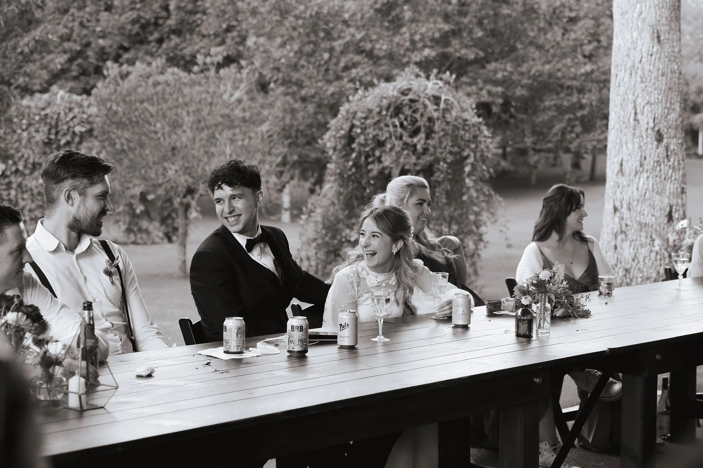 A group of people sitting at a long outdoor table, smiling and enjoying drinks, with trees in the background, during an outdoor gathering or celebration. Wedding Photography Waikato New Zealand. Wedding Photography Hamilton New Zealand