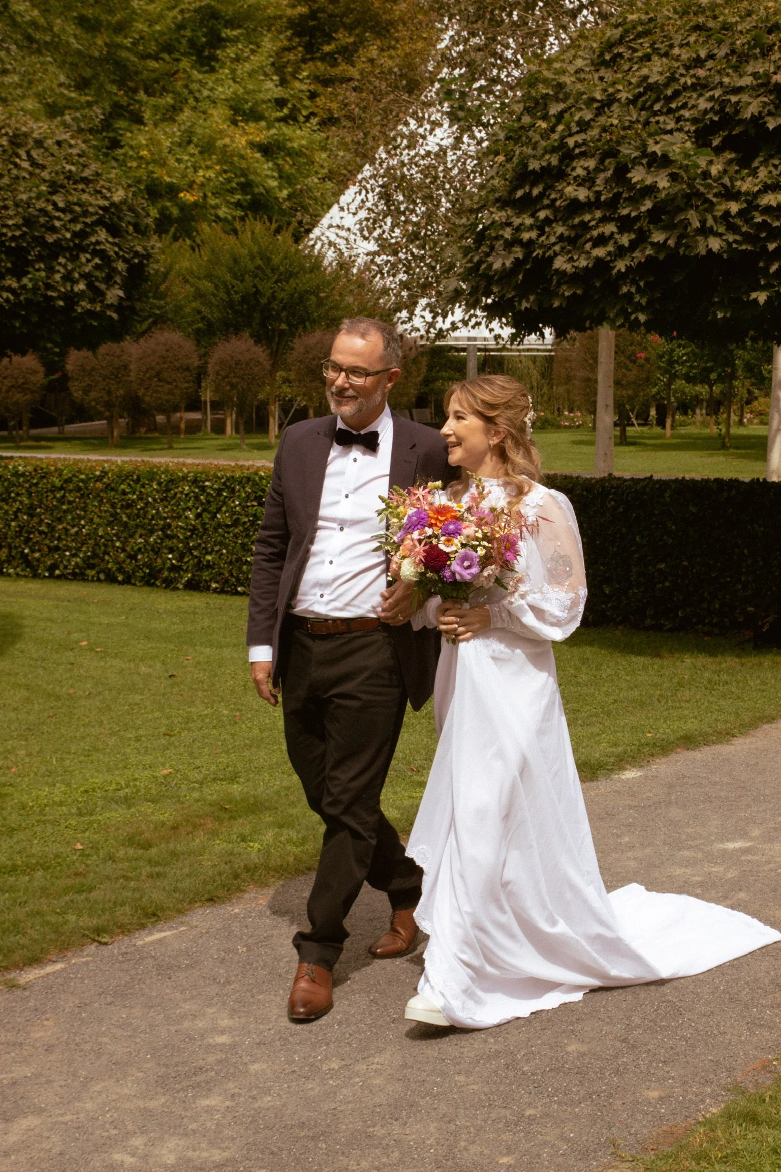 A bride in a white wedding dress holding a colorful bouquet of flowers walking arm-in-arm with a groom in a black tuxedo with a bow tie in a park with green trees and hedges. Wedding Photography Waikato New Zealand. Wedding Photography Hamilton 