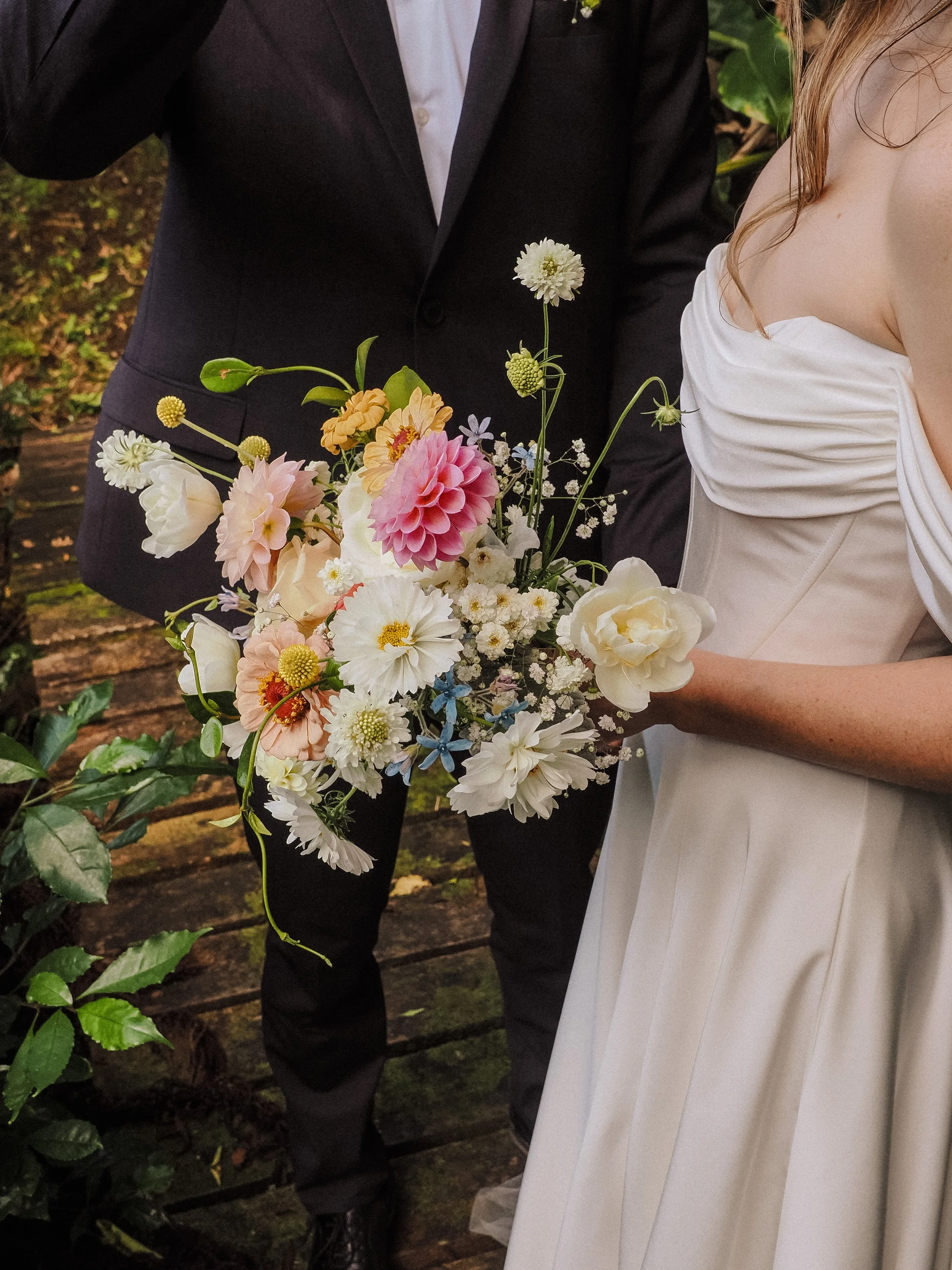 Close-up of a bride holding a colorful bouquet of flowers, with a groom partly visible in a black suit in the background.  Wedding Photography Waikato New Zealand. Wedding Photography Hamilton New Zealand. Northland Wedding Photography