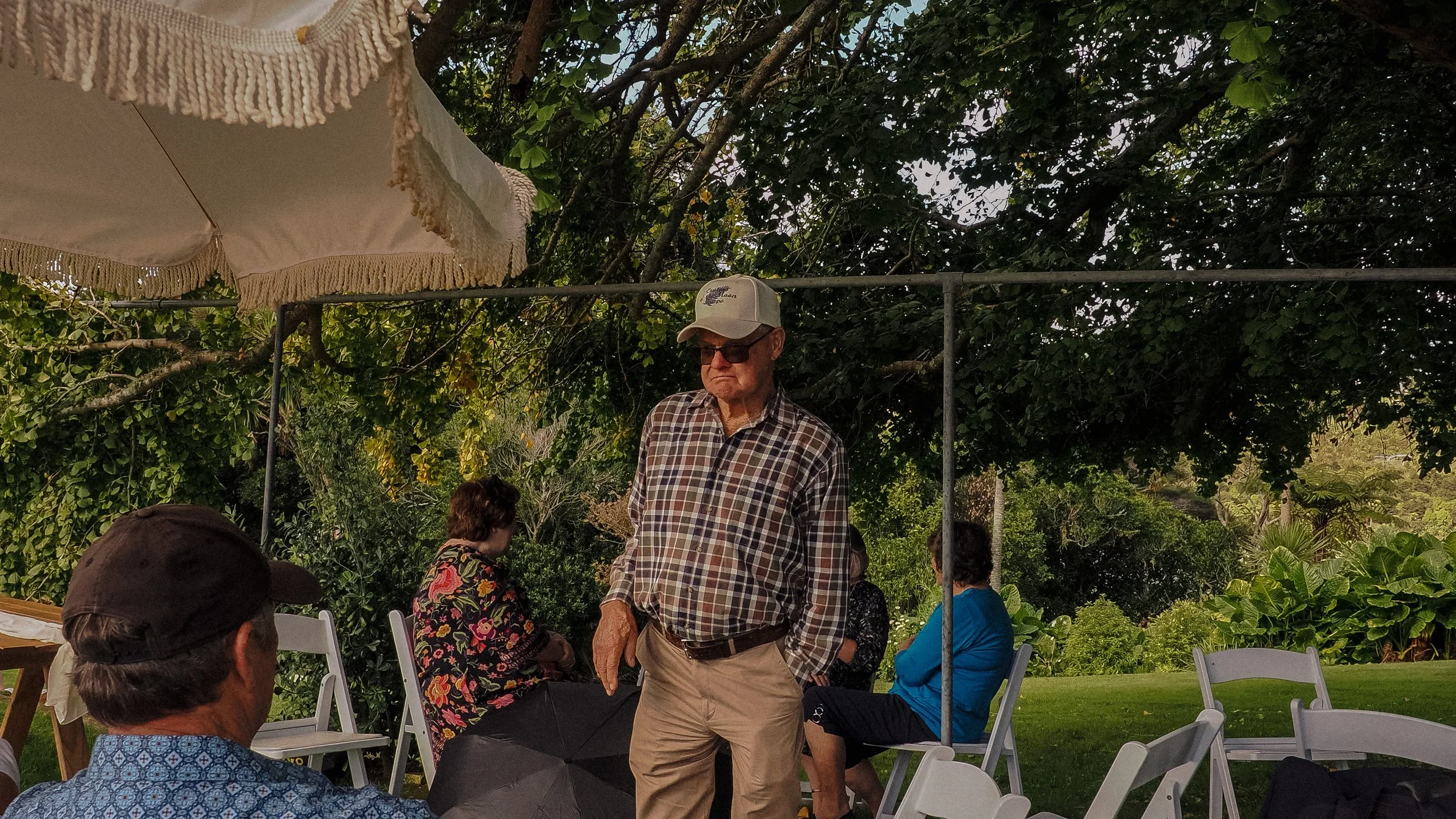 Older man wearing a plaid shirt, beige pants, sunglasses, and a white baseball cap standing outdoors under a tree canopy with people sitting and standing around him, some on white folding chairs, near a garden or park area.  Wedding Photography 