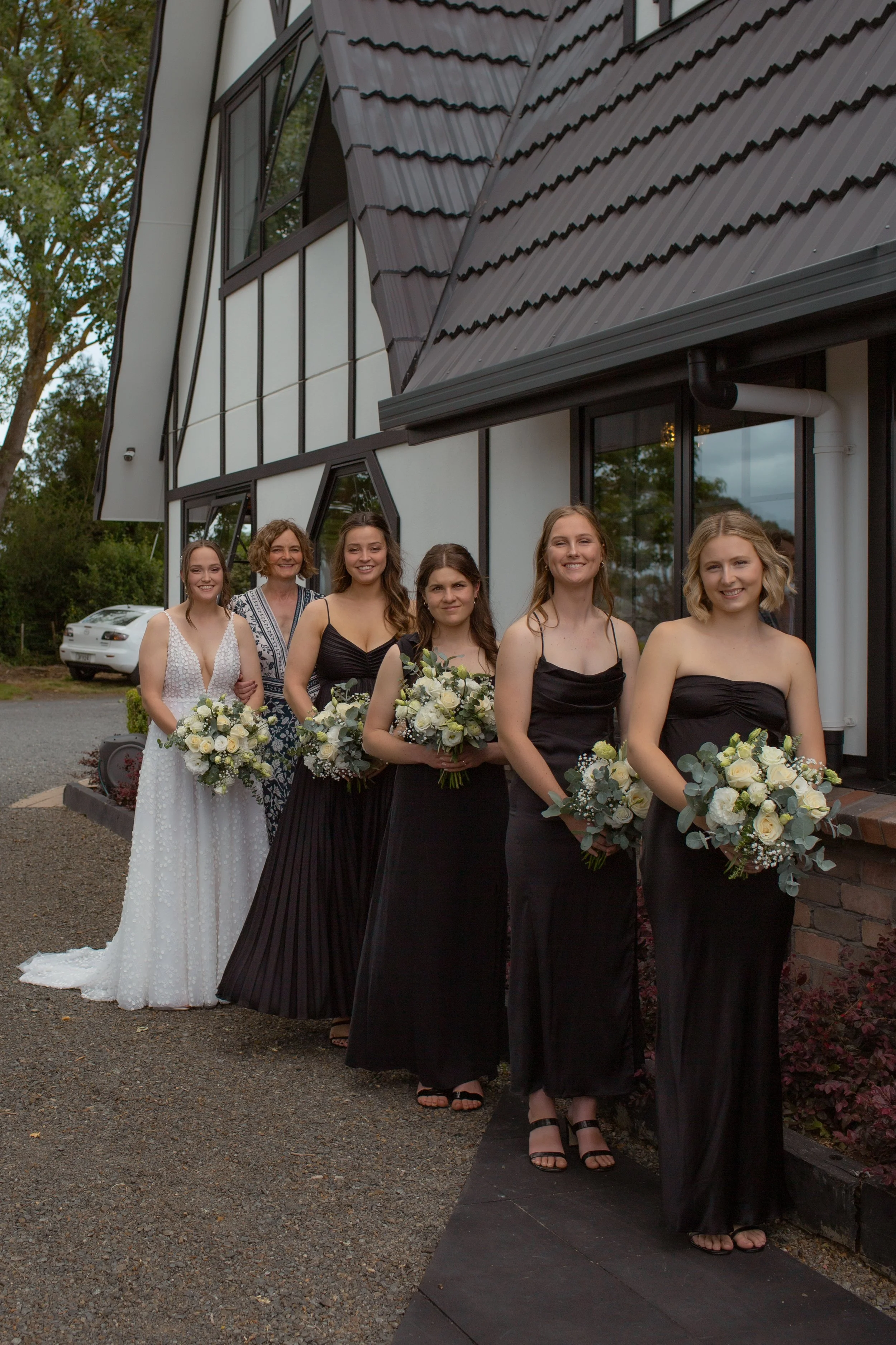 Bridesmaids and a bride standing outside a modern building, holding bouquets of white and green flowers, dressed in elegant black dresses, with a woman in a white dress at the back, all smiling.  Wedding Photography Waikato New Zealand. Wedding Photo