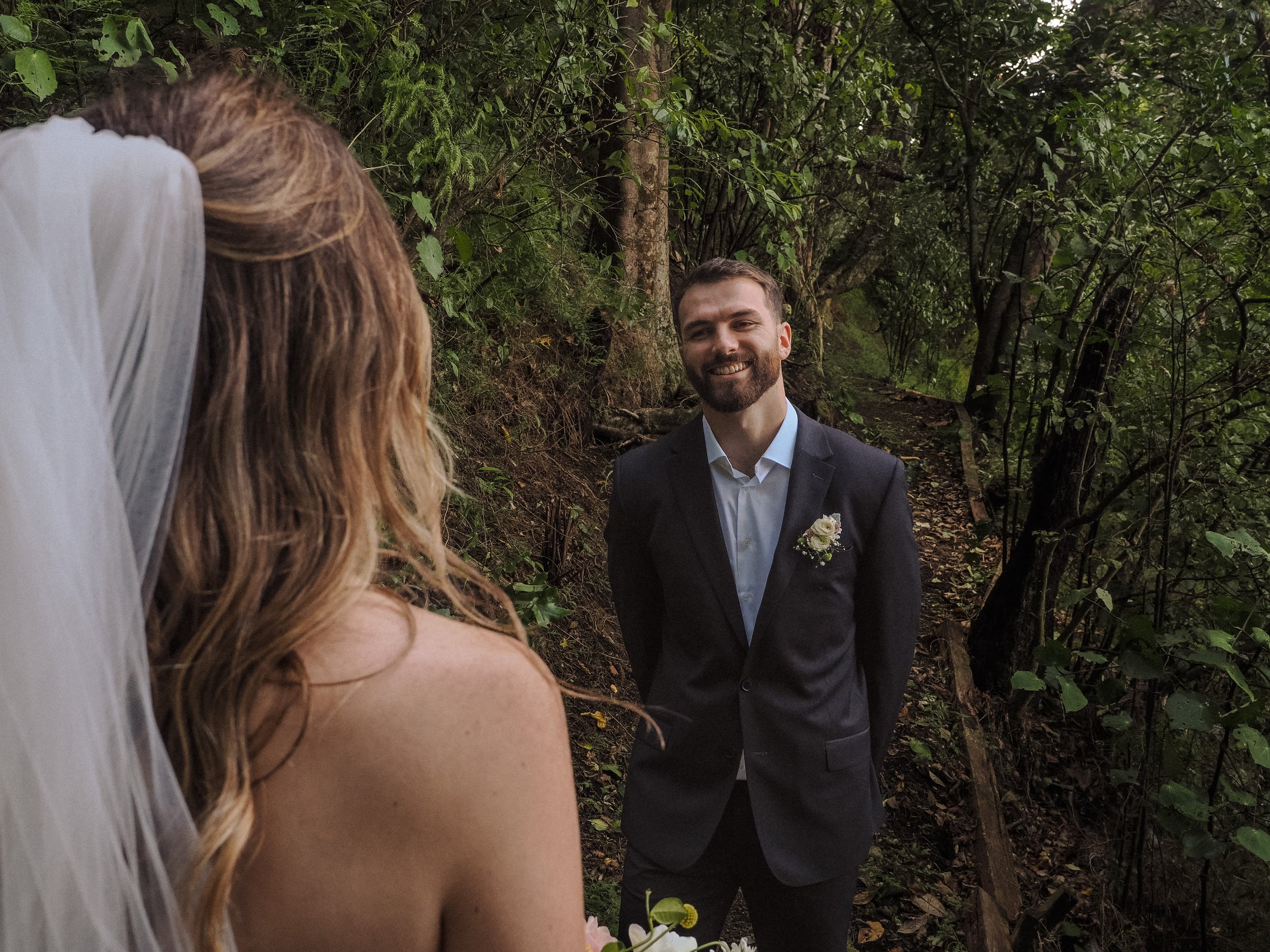 A bride and groom are seen in a forest during their wedding photos, with the groom dressed in a dark suit and the bride wearing a white veil and dress. The groom is smiling at the bride who is partly visible in the foreground.