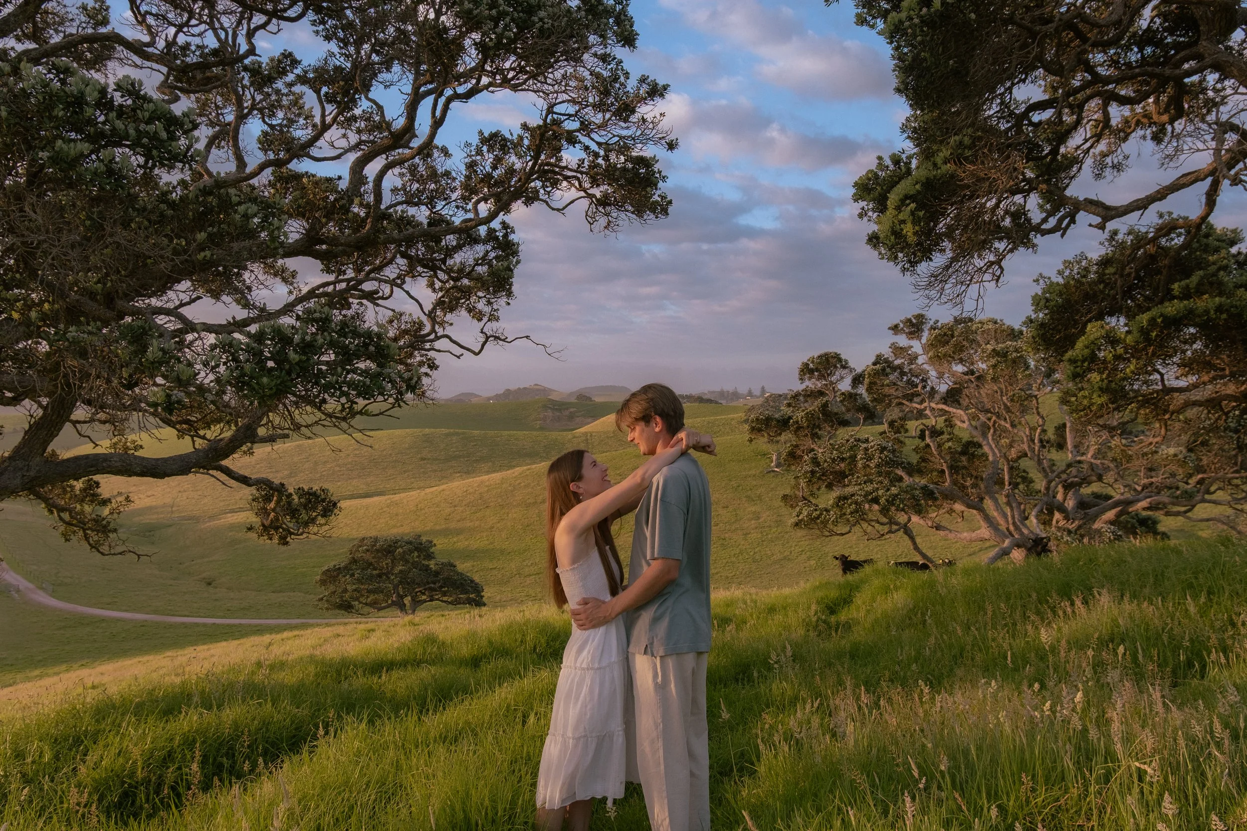 A young couple embracing outdoors in a lush green landscape with rolling hills and large trees under a partly cloudy sky, during sunset or late afternoon.
