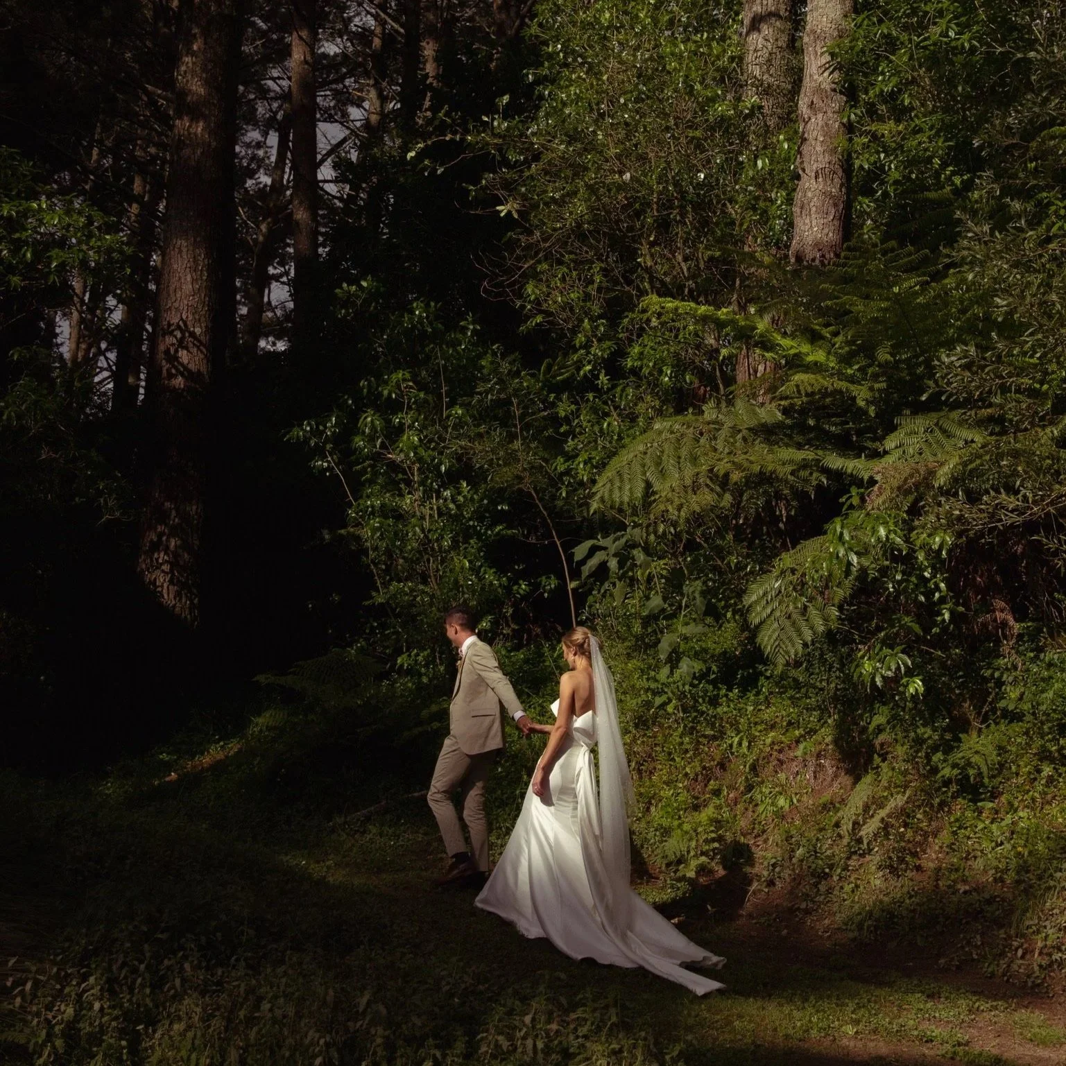 A bride and groom holding hands and walking through a lush, green, wooded area at sunset or dusk, with the bride in a white wedding dress and the groom in a tan suit.