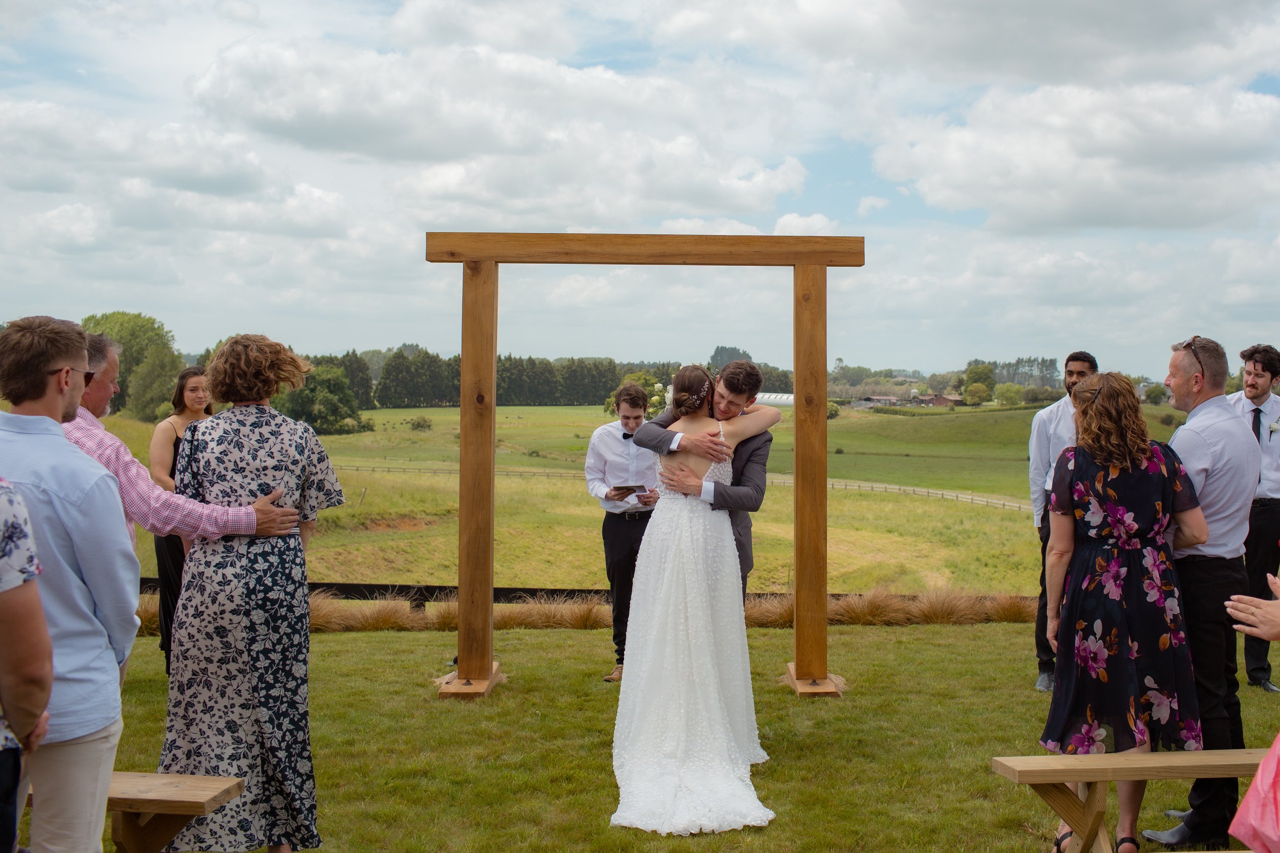 A couple gets married outdoors under a wooden arch, hugging as the bride wears a white gown and the groom in a gray suit, surrounded by friends and family on a grassy field with a rural landscape in the background.