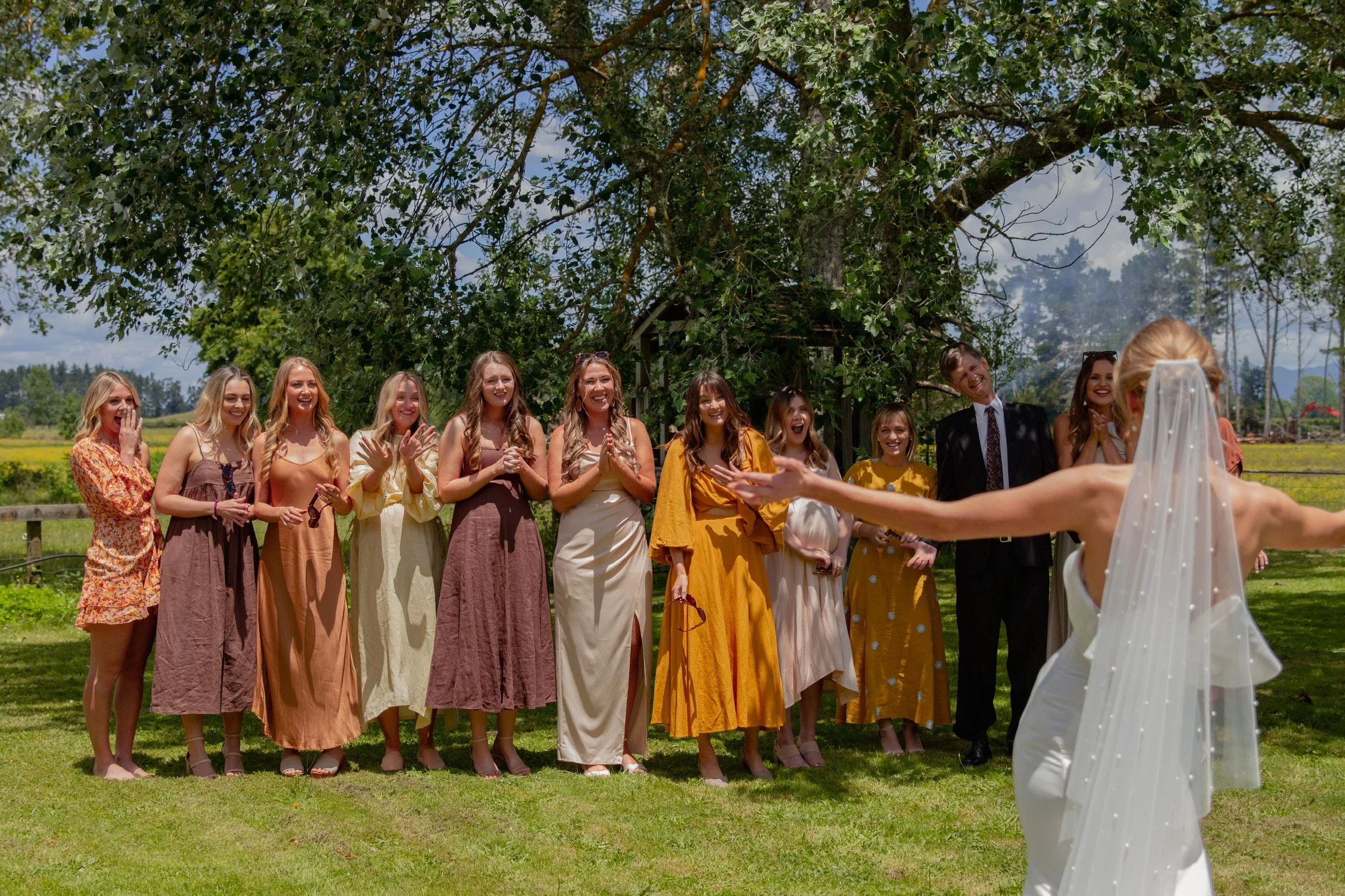 A bride in a white wedding dress and veil stands outdoors with arms outstretched, facing a group of women and one man under a large tree, during a wedding celebration.  Wedding Photography Waikato New Zealand. Wedding Photography Hamilton New Zealand