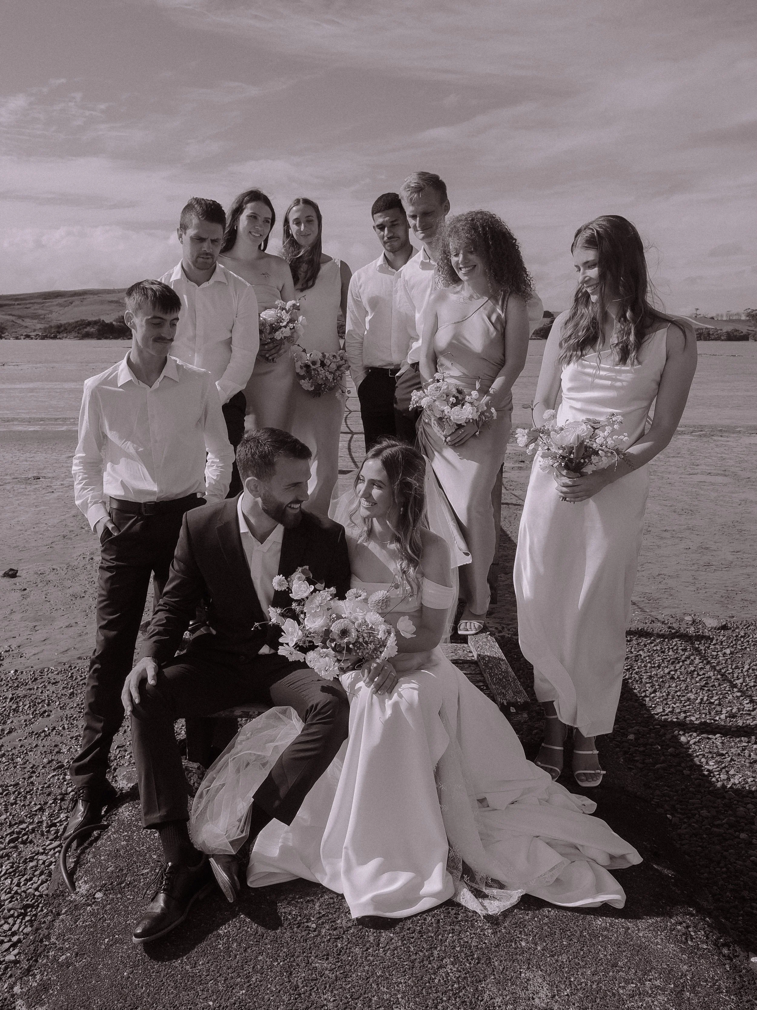 A group of wedding friends, dressed in formal attire, gathered outdoors near a beach, with the bride and groom sitting in front, surrounded by bridesmaids and groomsmen.  Wedding Photography Waikato New Zealand. Wedding Photography Hamilton 