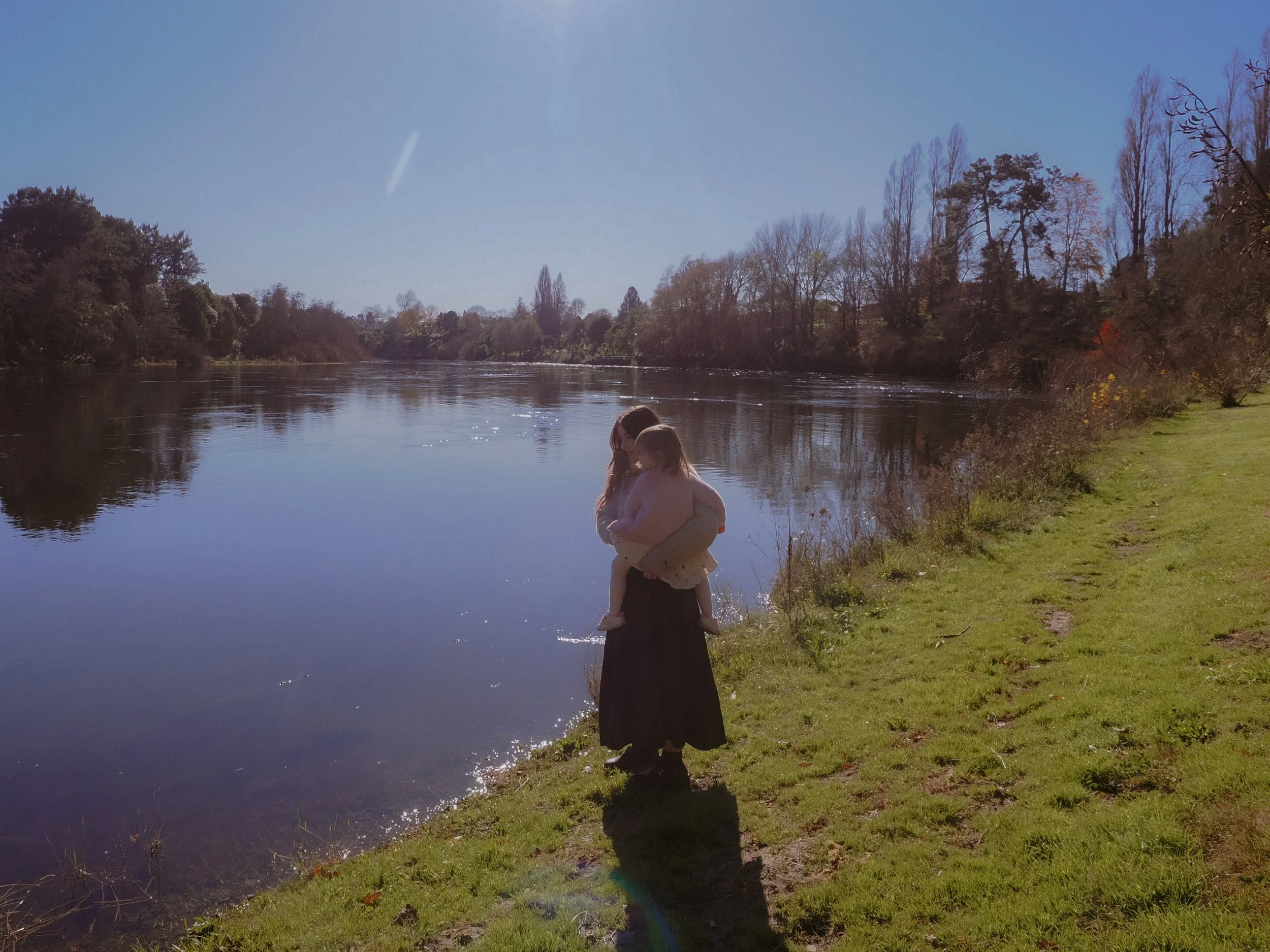 Woman holding a child by a river on a sunny day with trees on the opposite bank and a clear blue sky. Family Photography Waikato New Zealand. Family Photography Hamilton New Zealand. Northland Wedding Photography