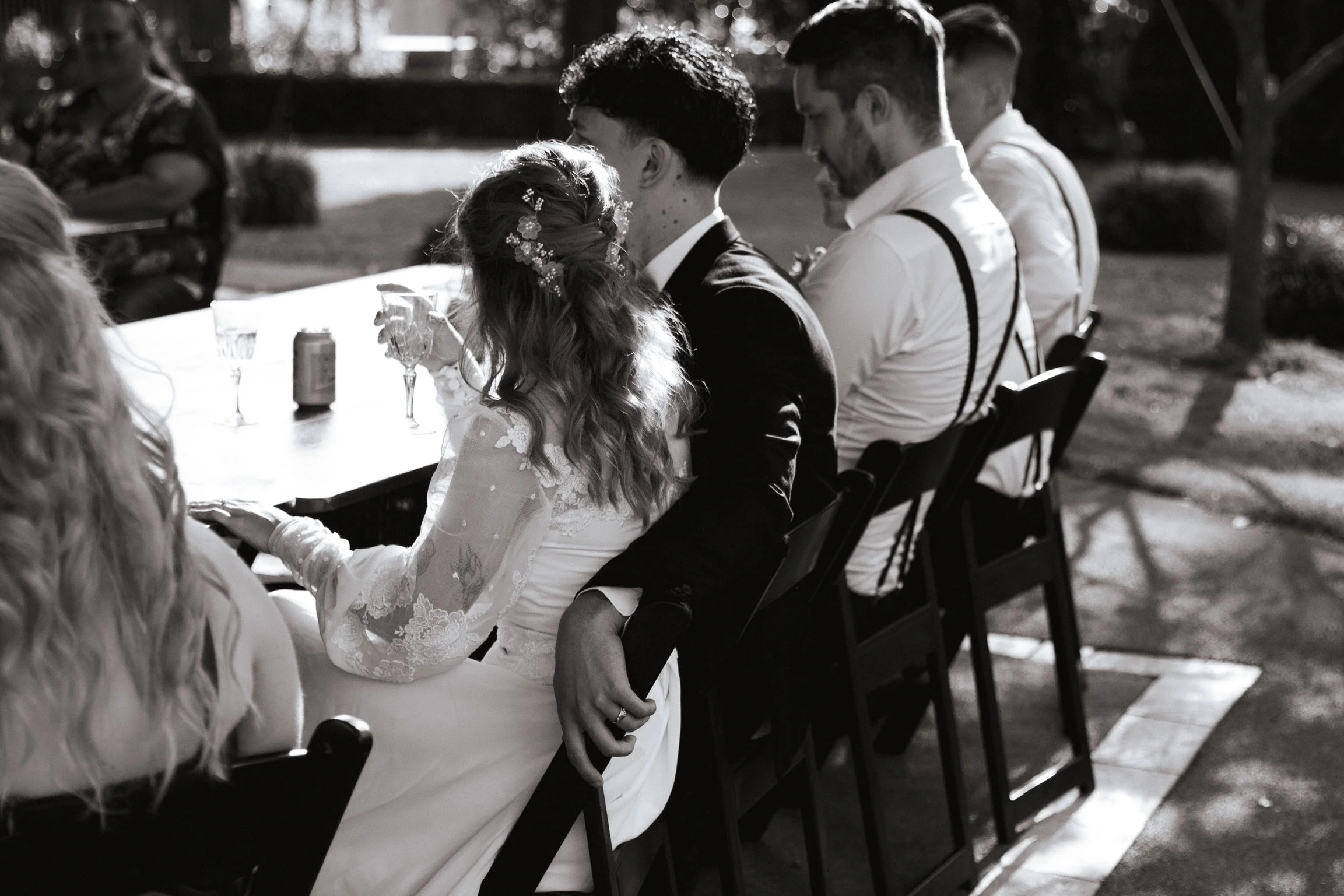 People sitting at a table outdoors, dressed in wedding attire, sharing a tender moment during a wedding reception. Wedding Photography Waikato New Zealand. Wedding Photography Hamilton New Zealand