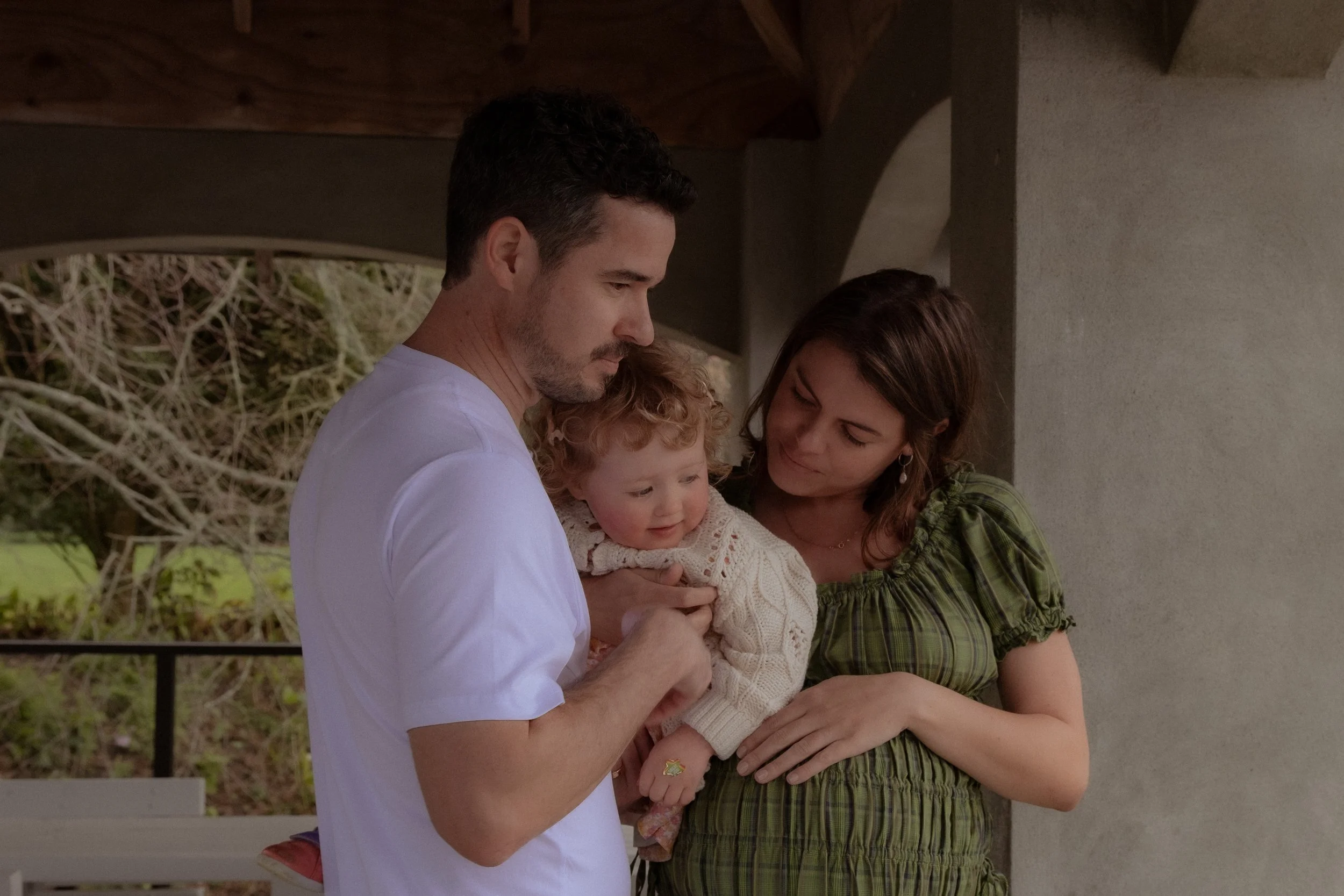 A man, woman, and young girl are gathered closely together outside, sharing a tender moment. The man holds the girl while the woman looks on warmly. They are standing near a building with columns and greenery in the background.