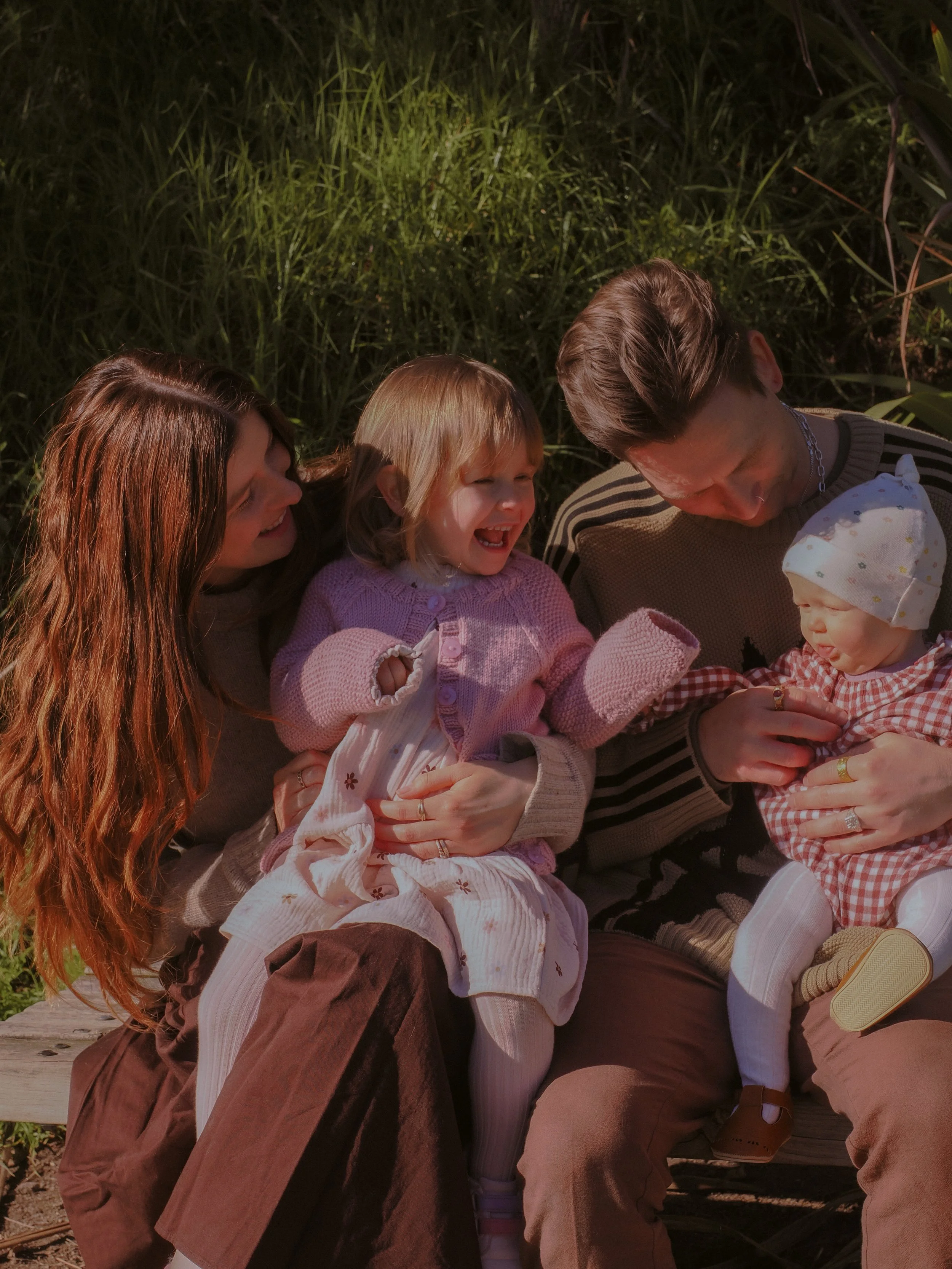 A family of four, including a mother, father, young girl, and baby girl, sitting outdoors on a wooden bench, enjoying a moment together in a sunny, grassy area. Family Photography Waikato New Zealand. Family Photography Hamilton New Zealand. 