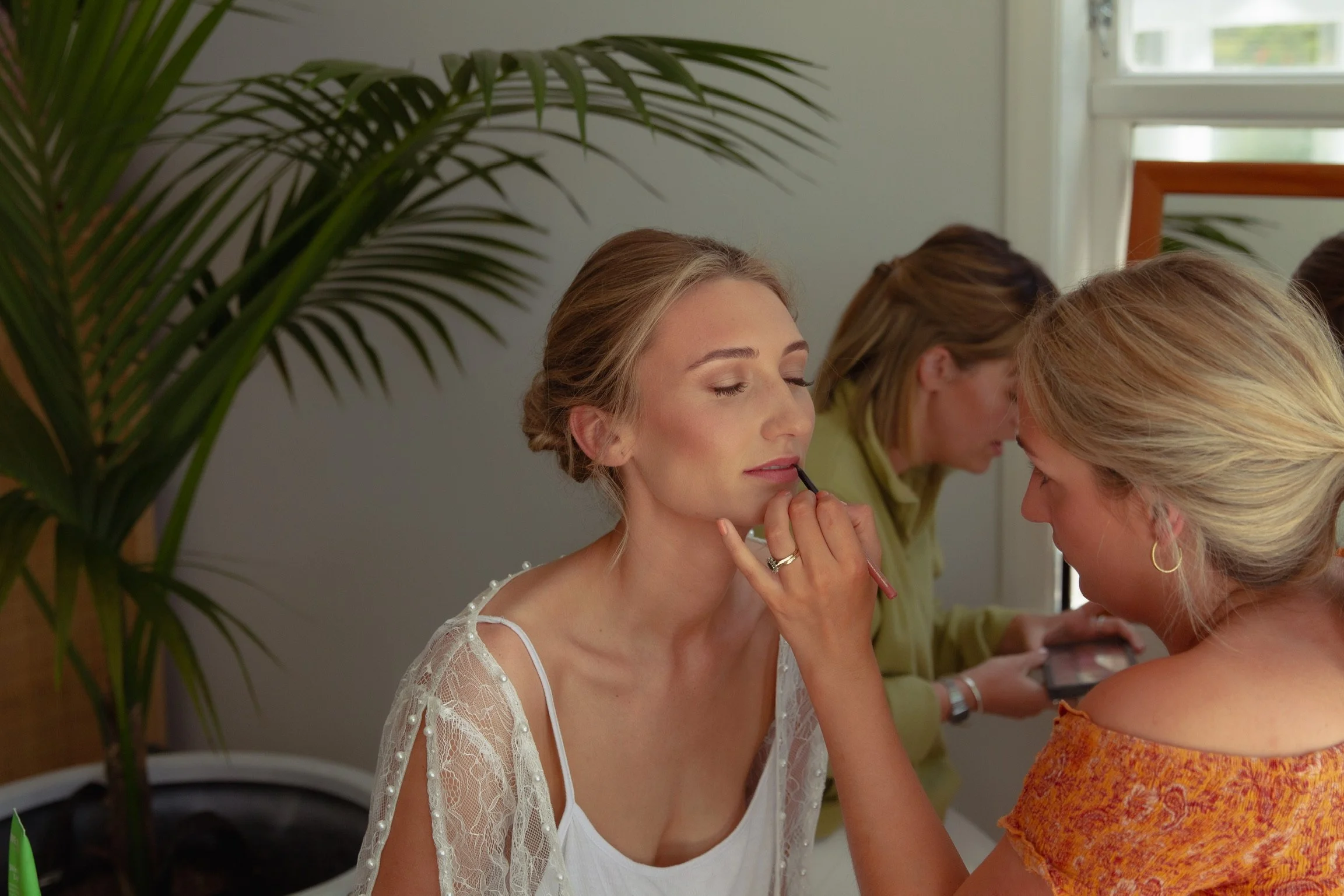 A woman with light brown hair in an elegant updo has her makeup applied by a makeup artist in a light orange off-shoulder top. There is a large green plant and a woman in the background using a phone.