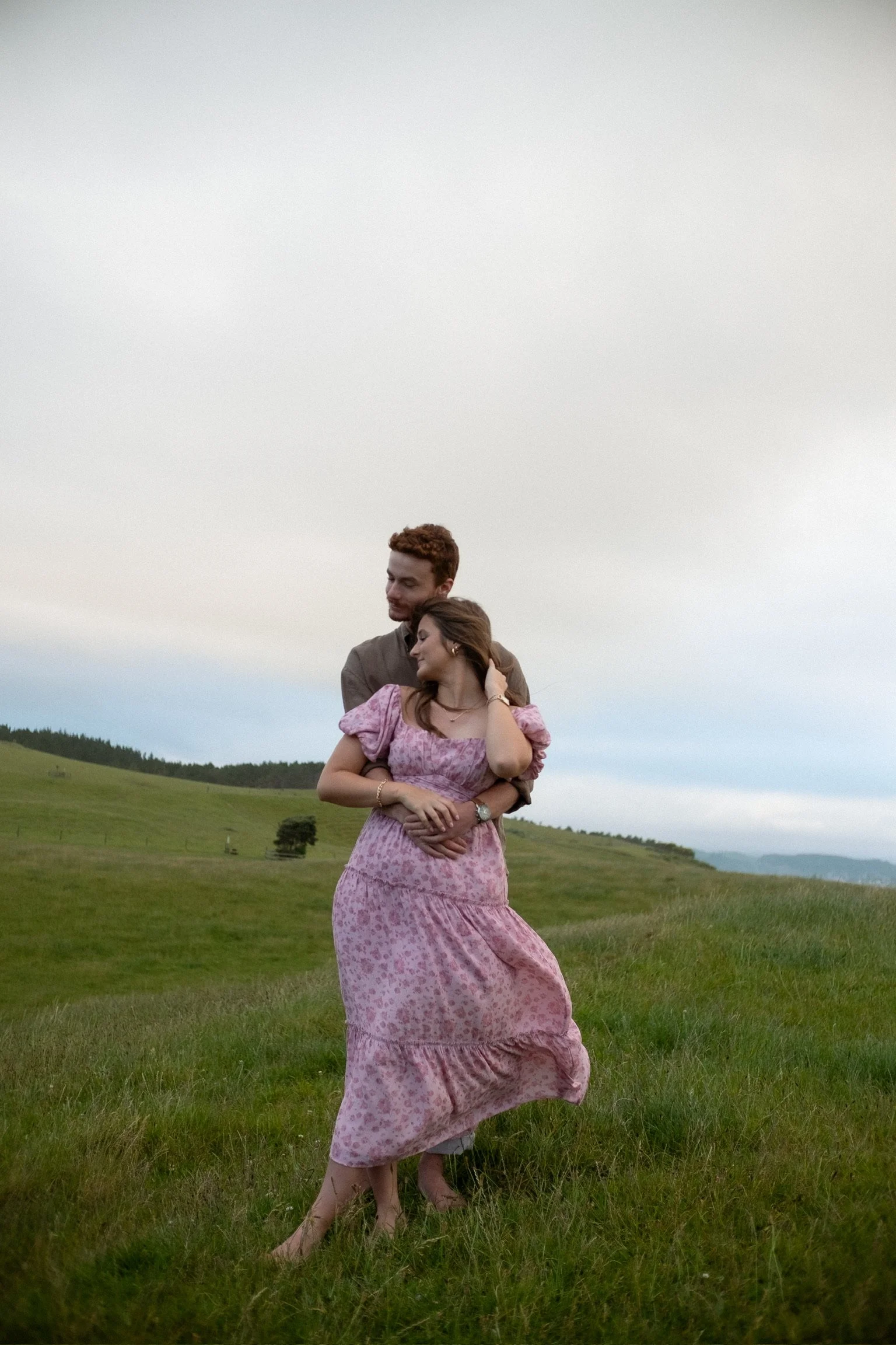 A couple embracing in a grassy field during daytime, with a cloudy sky in the background. Couples photography Raglan New Zealand. Engagement photography Hamilton New Zealand. Whangarei New Zealand Couples photographer