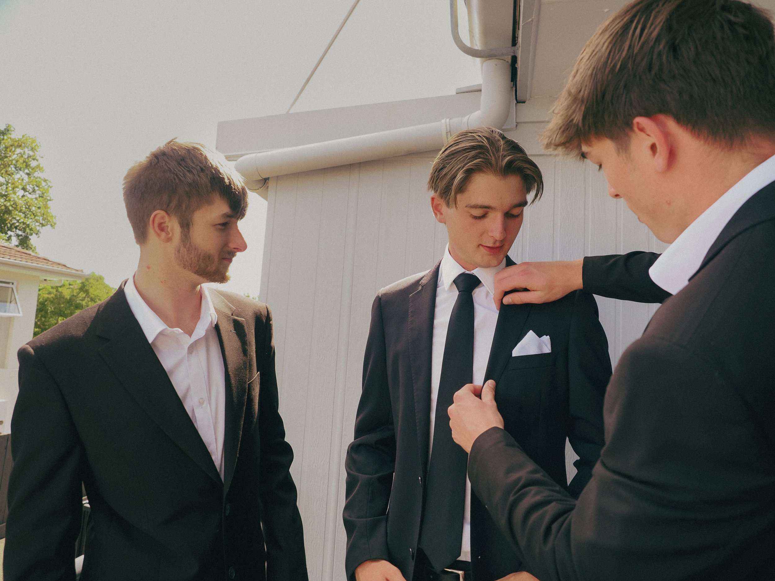 Four young men dressed in suits are gathered outside, with one man adjusting another man's jacket or tie. They appear to be preparing for a formal event or ceremony.
