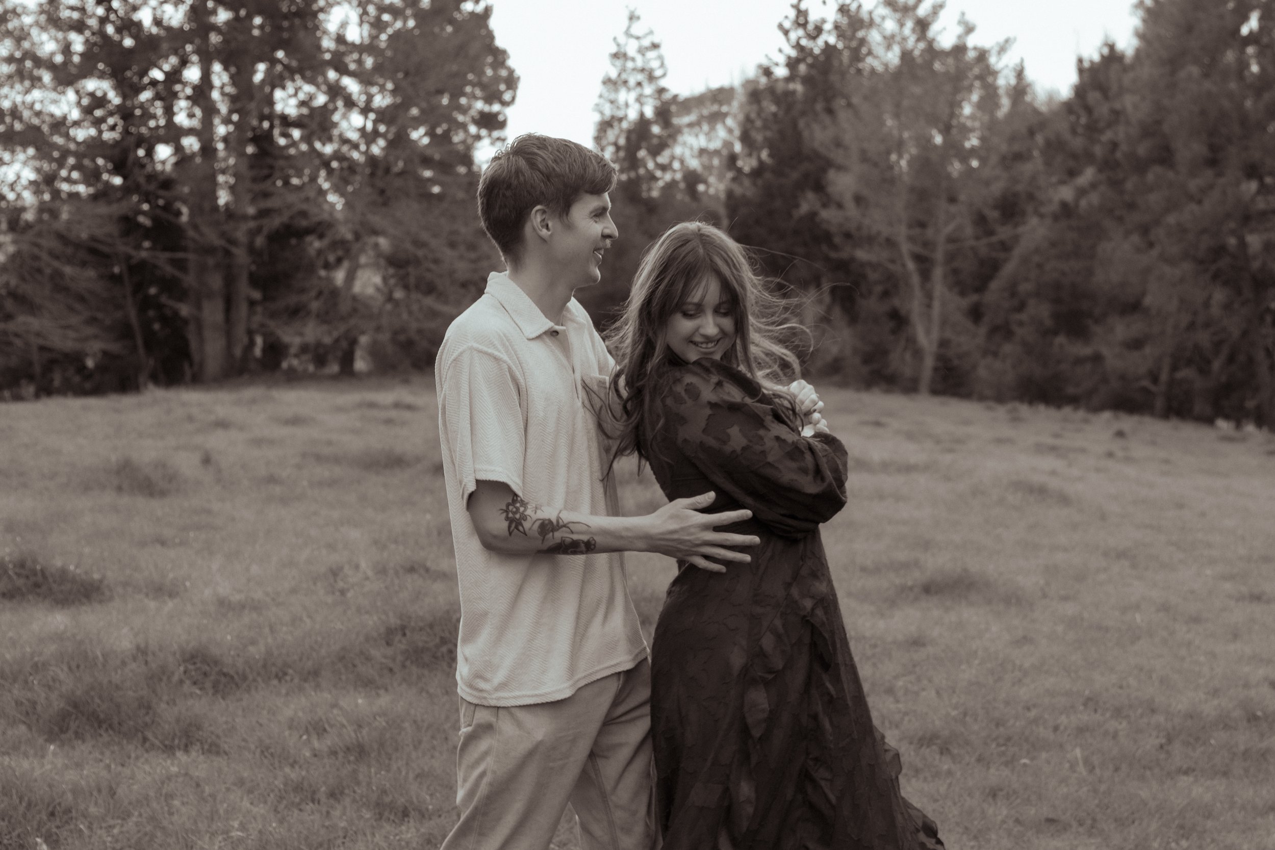 A man and woman are smiling and laughing outdoors in a grassy field with trees in the background. Couples photography Hamilton New Zealand. Engagement photography Hamilton New Zealand. Whangarei New Zealand Couples photographer