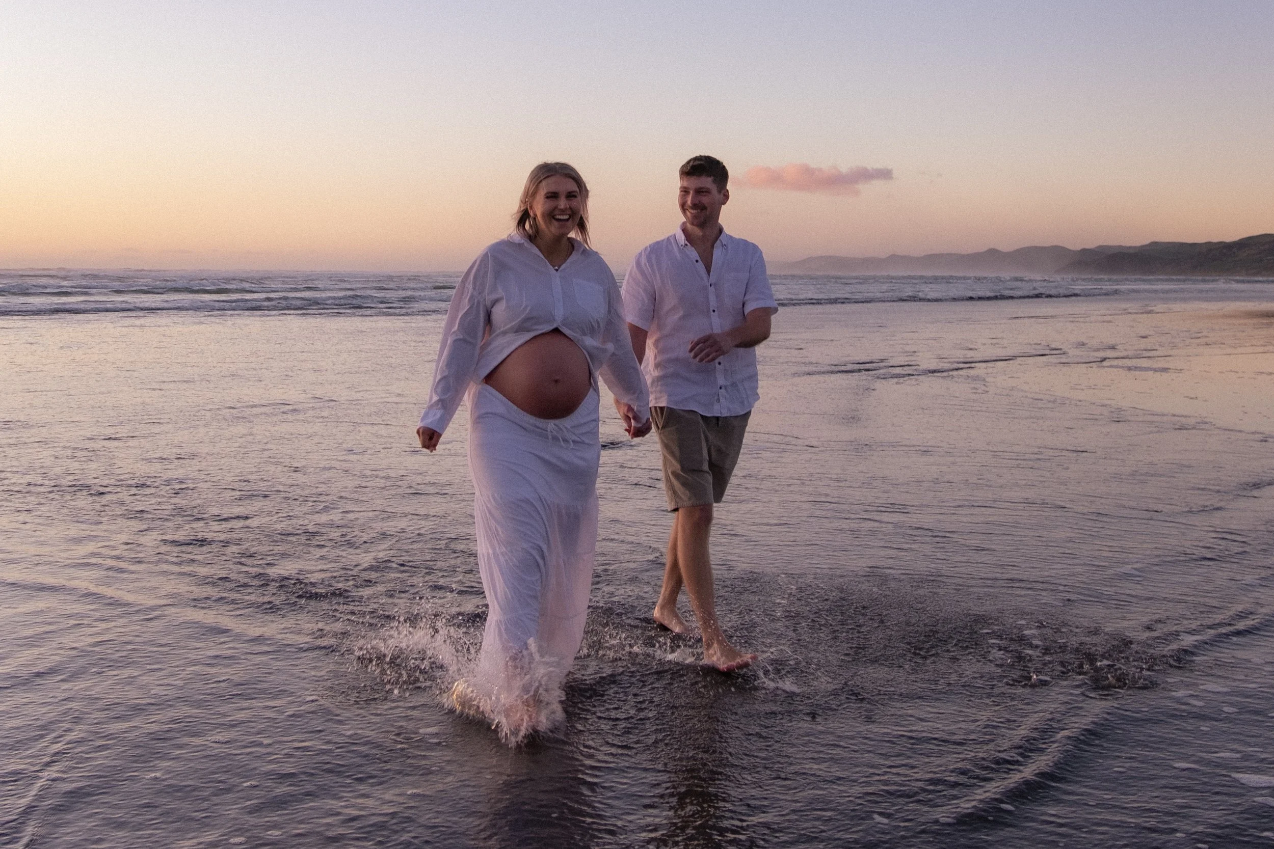 A pregnant woman and a man walking on the beach at sunset, holding hands and smiling, with the woman wearing a white wrap-around skirt and an unbuttoned white shirt, and the man wearing a white shirt and beige shorts. Maternity photography 