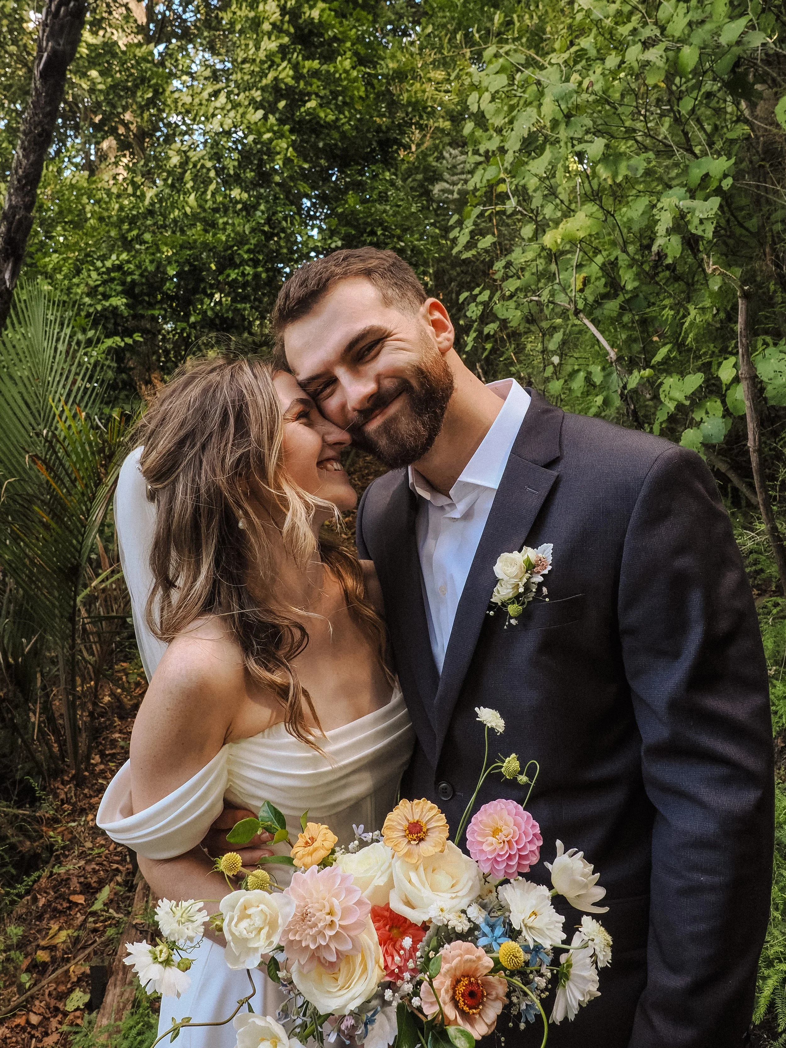 A newlywed couple, a woman with long wavy hair in a white wedding dress and a man with short hair and beard in a black suit, hugging in a lush green outdoor setting, holding a colorful bouquet of flowers.  Wedding Photography Waikato New Zealand.