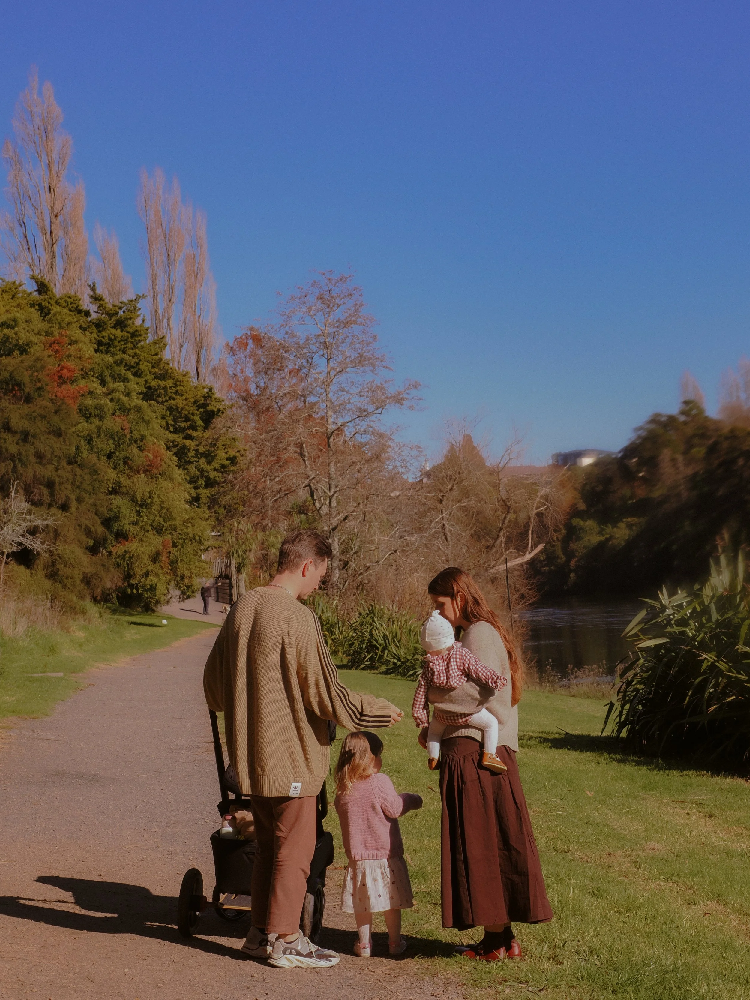 A family of four, including a man, woman, and two children, enjoying a day outside by a river, with trees and clear blue sky in the background. Family Photography Waikato New Zealand. Family Photography Hamilton New Zealand. Northland Wedding Photo