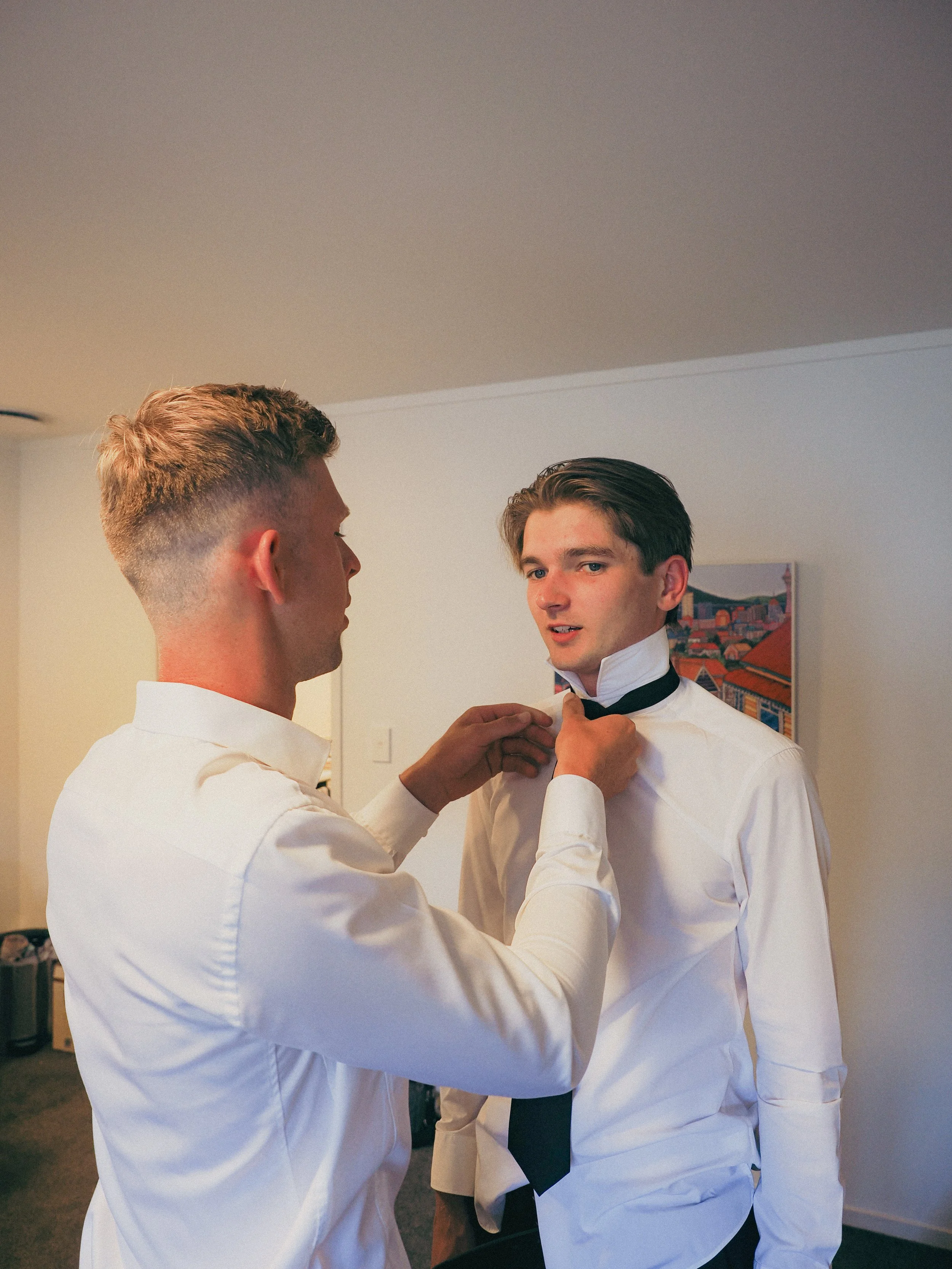 Two young men dressed in white shirts with black ties, one is helping the other adjust his tie in a room with white walls and a colorful painting in the background.