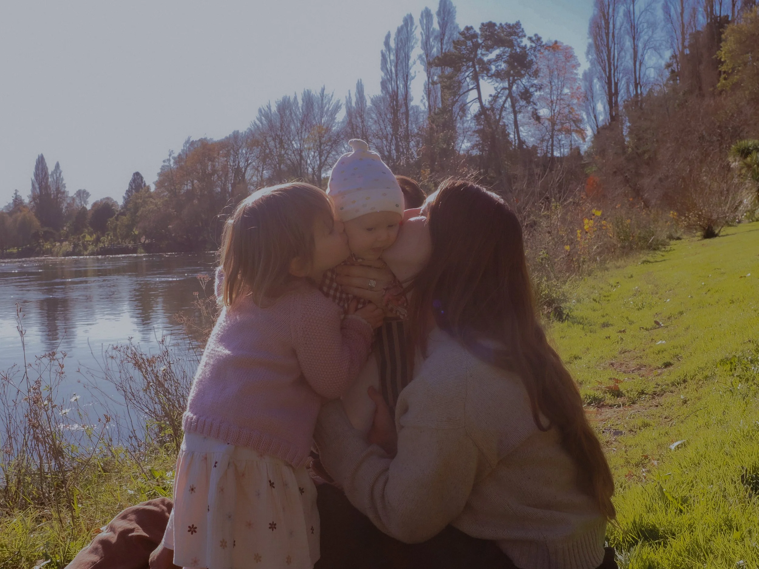 Two young girls, one with long dark hair and the other with shoulder-length hair, are giving a baby a kiss on the cheeks outdoors near a body of water with trees in the background. The baby is wearing a white hat with yellow polka dots and is being h