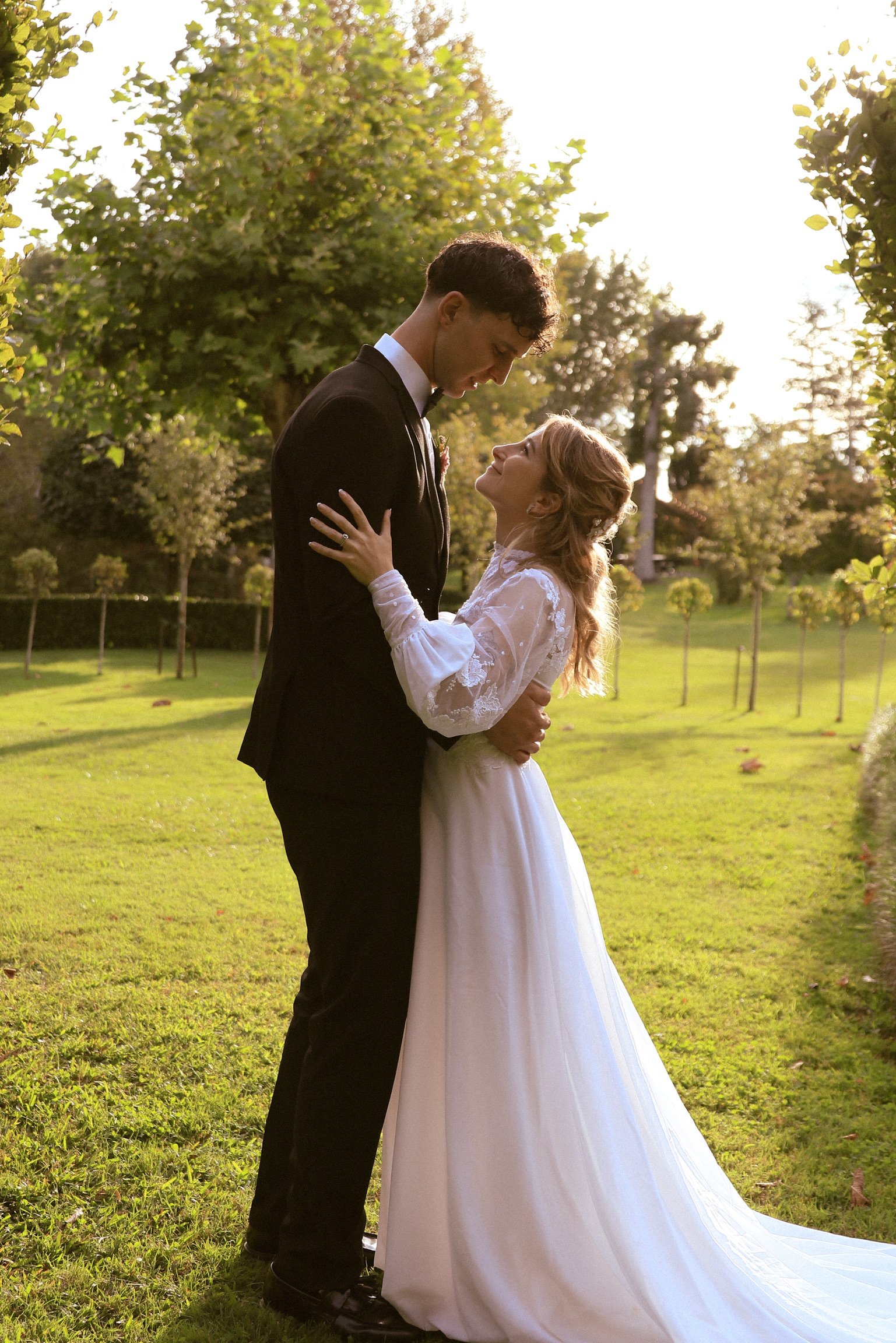 A couple in wedding attire standing close to each other outdoors during sunset. The groom in a black tuxedo and the bride in a white lace gown are gazing into each other's eyes with greenery and trees in the background.