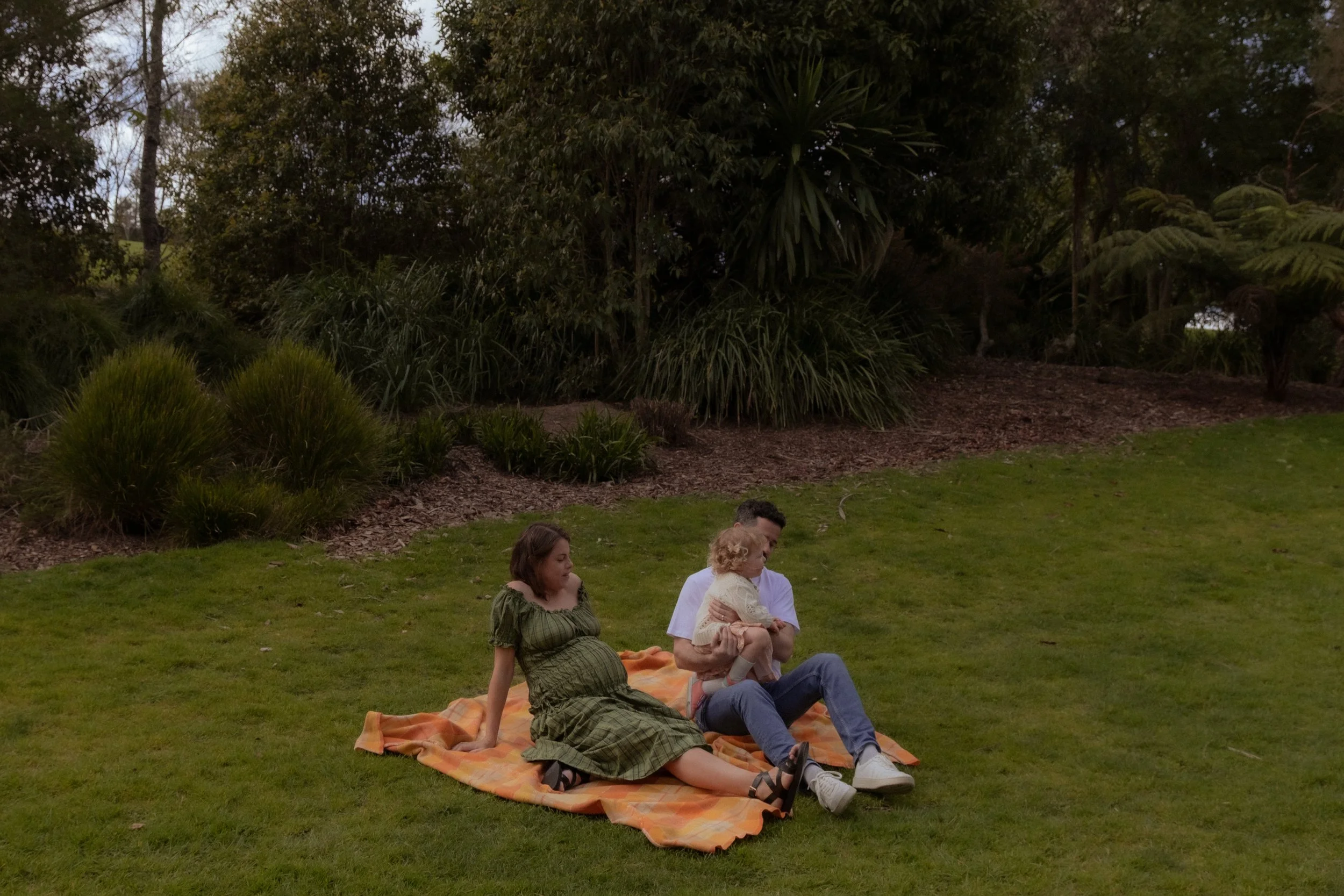 Family of three sitting on a blanket in a park with lush trees and bushes in the background. Family Photography, Maternity photographer, Hamilton New Zealand Photographer