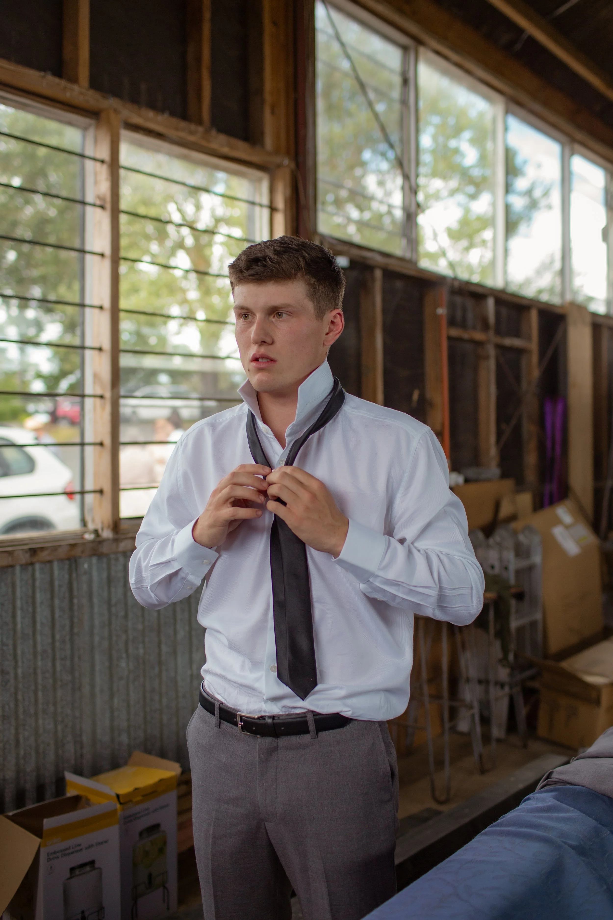 A young man in a white dress shirt and grey pants is adjusting a black tie in a rustic indoor space with large windows and wooden walls.  Wedding Photography Waikato New Zealand. Wedding Photography Hamilton New Zealand. Northland Wedding Photography