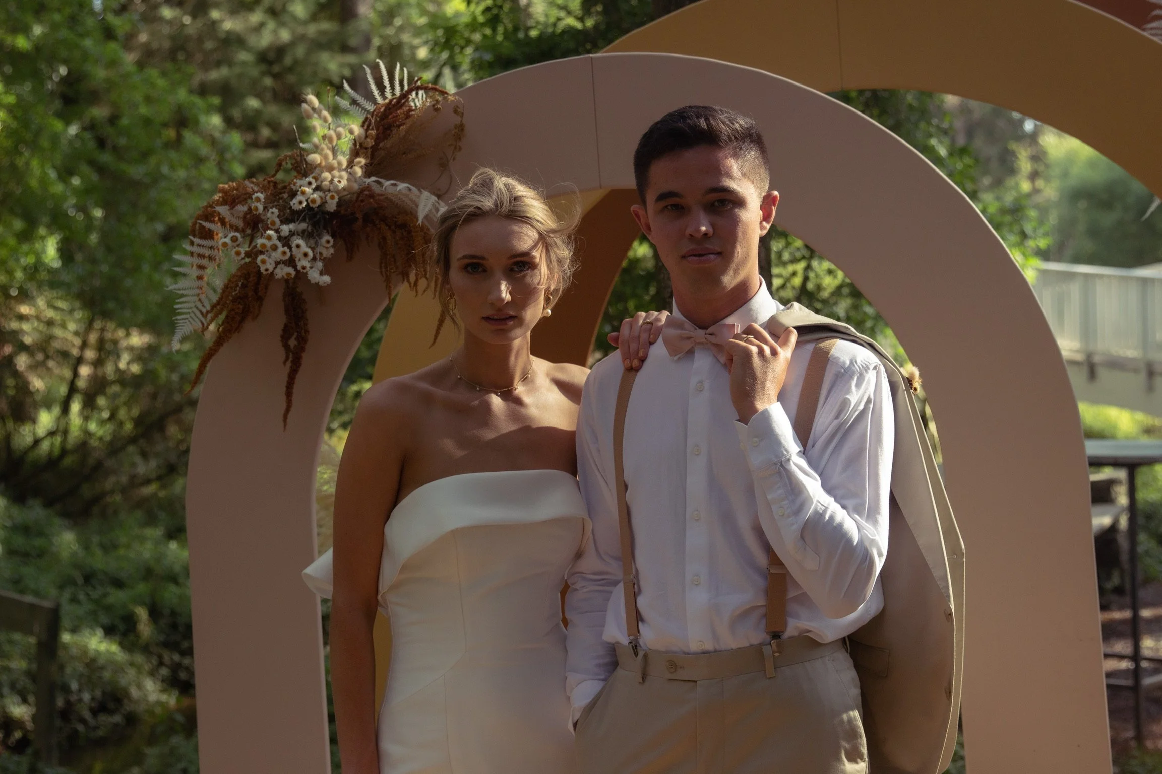 A bride and groom standing outdoors under a decorative arch with flowers, during a wedding celebration.  Wedding Photography Waikato New Zealand. Wedding Photography Hamilton New Zealand. Northland Wedding Photography