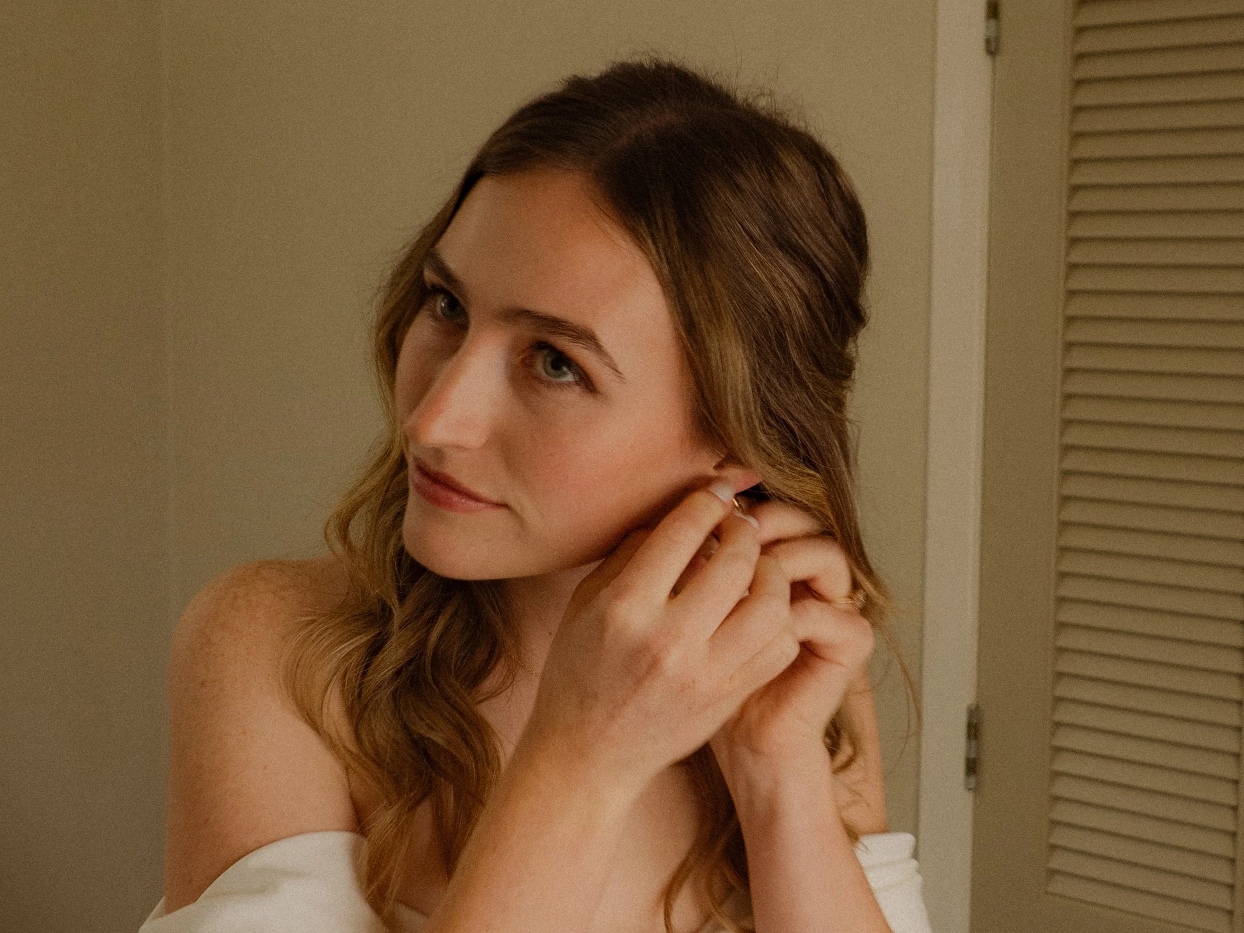 A young woman with wavy light brown hair is putting on earrings in front of a mirror.  Wedding Photography Waikato New Zealand. Wedding Photography Hamilton New Zealand. Northland Wedding Photography