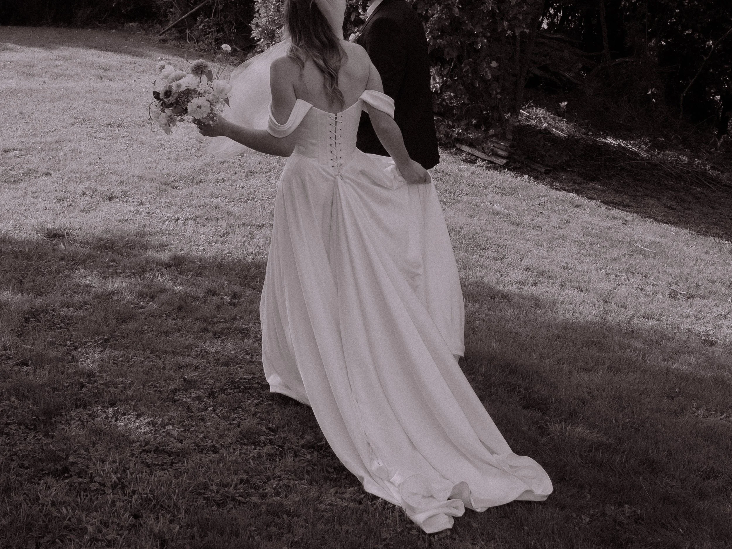 A bride in a strapless wedding gown holding a bouquet of flowers walks through a grassy outdoor area beside a person in dark clothing.
