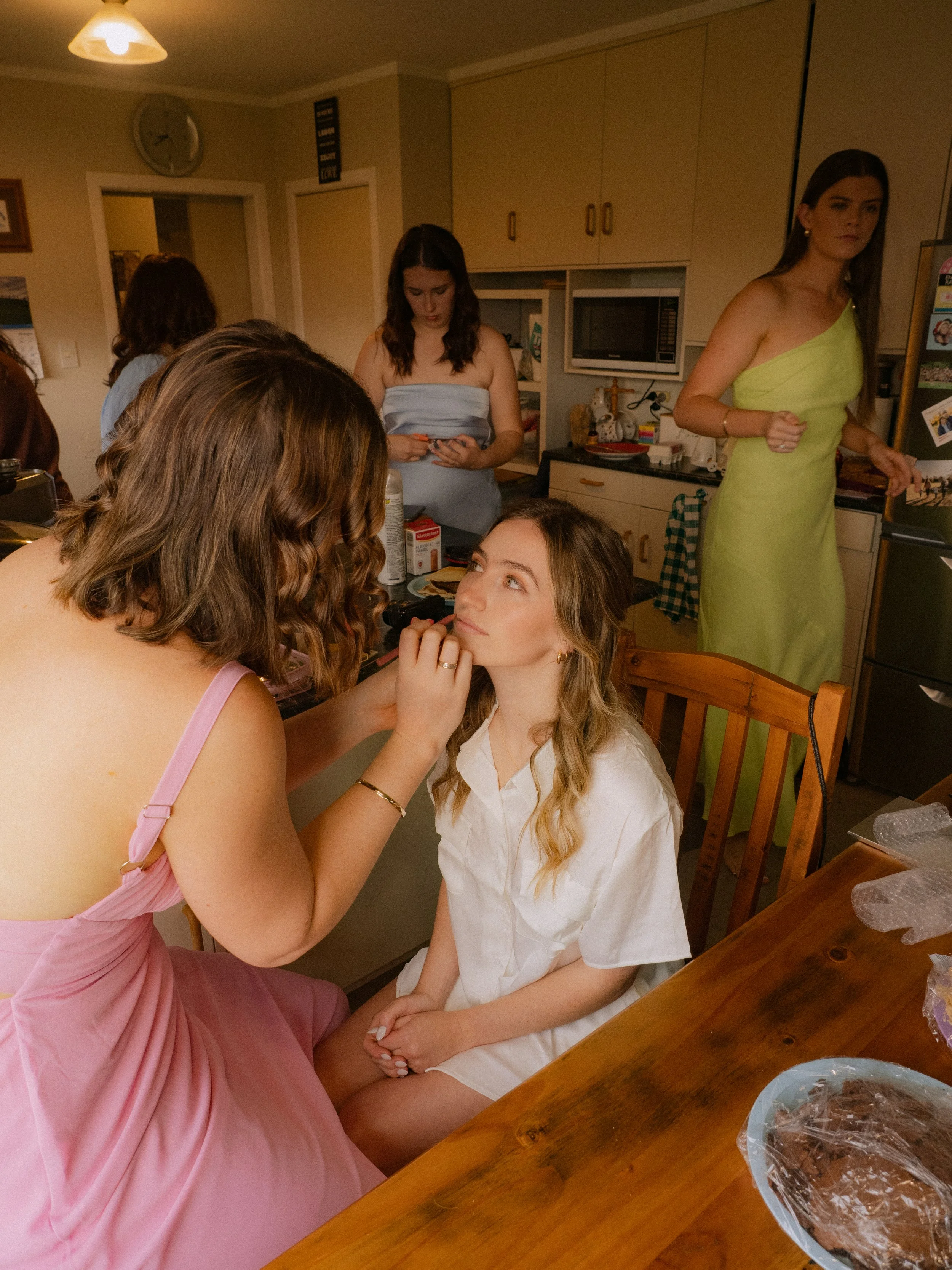 A woman in a pink dress applying makeup to a seated young woman in a white shirt in a kitchen, with other women preparing in the background.  Wedding Photography Waikato New Zealand. Wedding Photography Hamilton New Zealand. Northland Wedding Photo