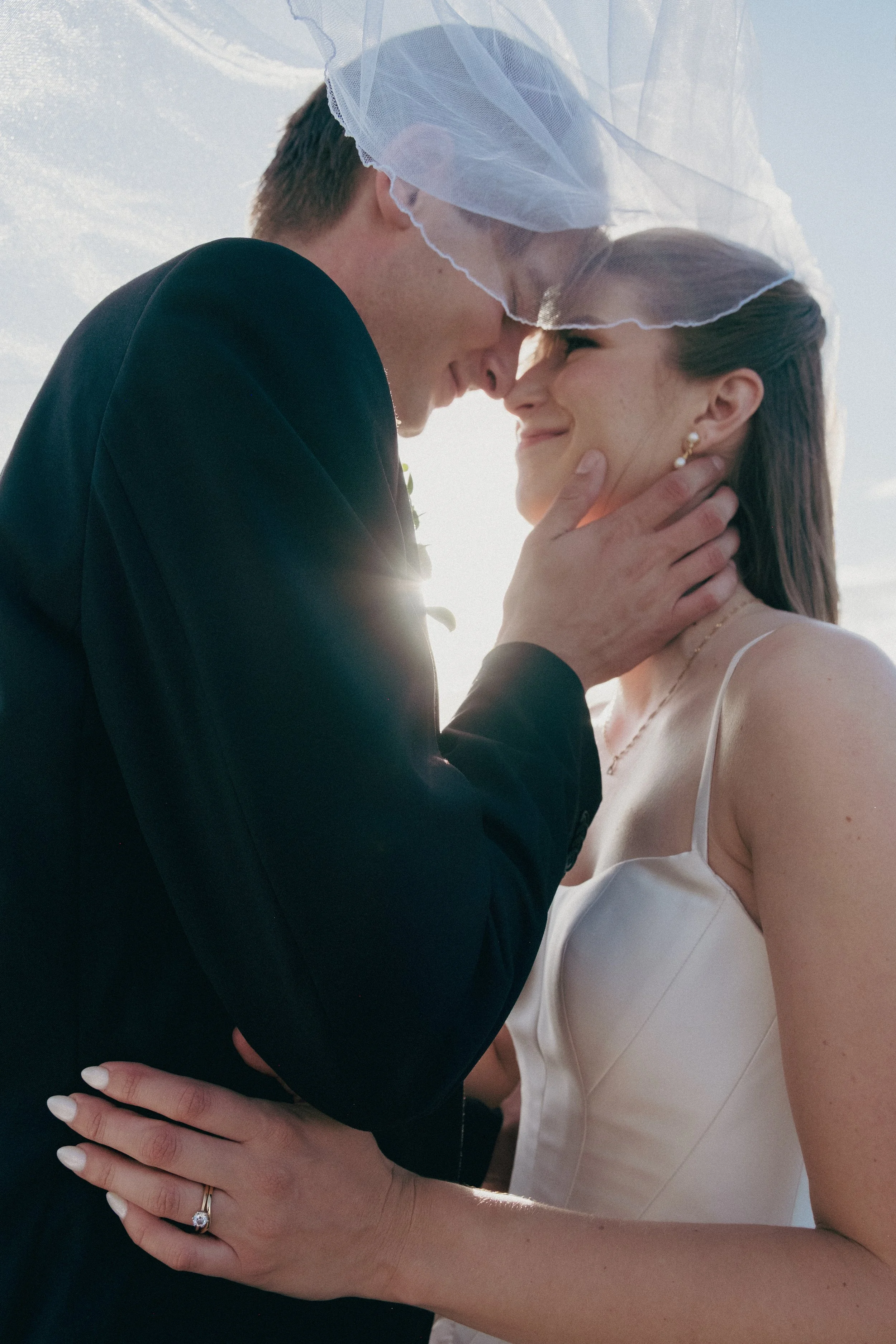 A bride and groom are touching foreheads and holding each other during their wedding ceremony, with the bride wearing a veil and white dress and the groom wearing a dark suit, under a bright sky.