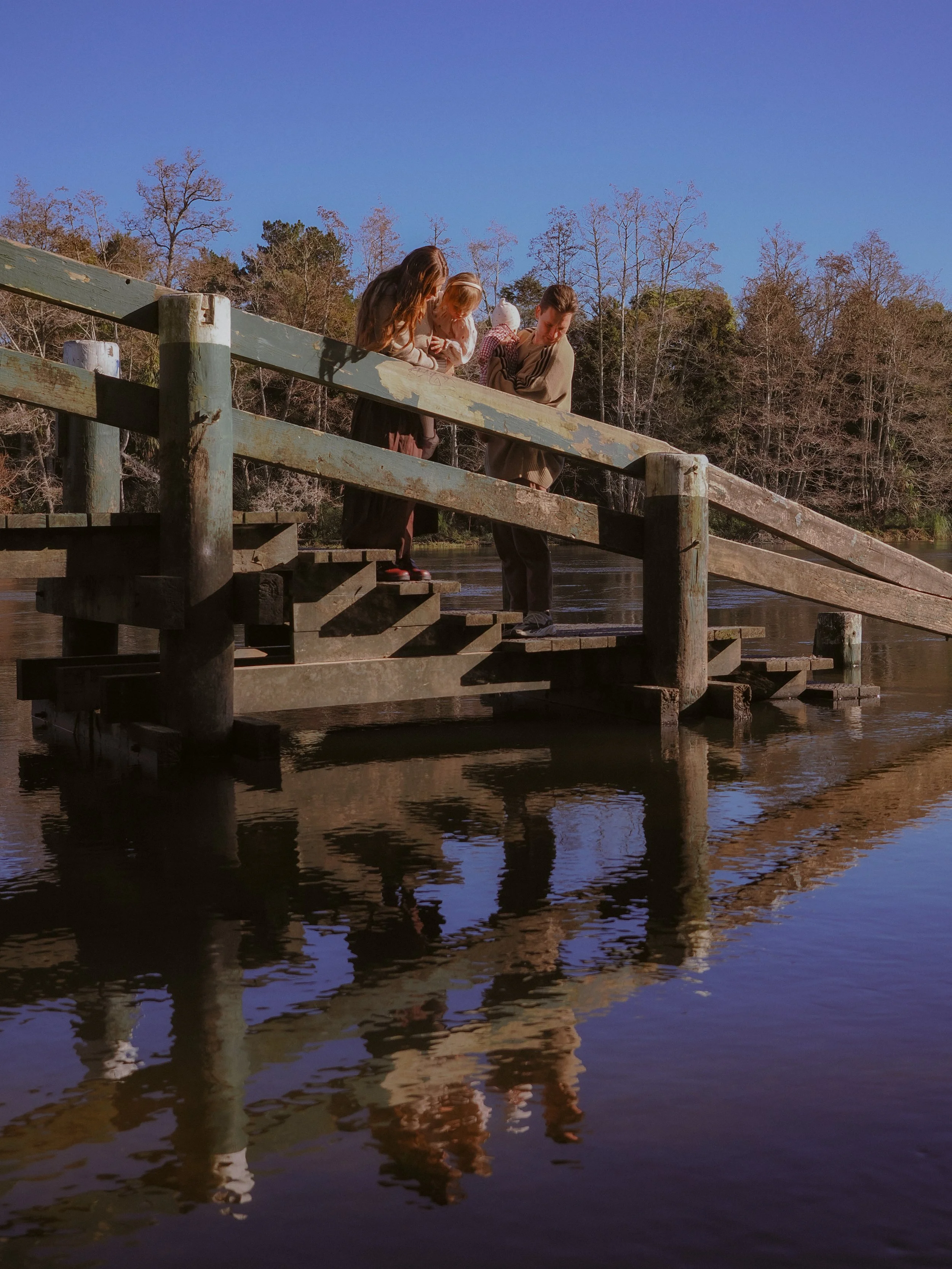 Three children and an adult standing on a wooden dock, looking at something in the water, with trees in the background on a clear day. Family Photography Waikato New Zealand. Family Photography Hamilton New Zealand. Northland Wedding Photography