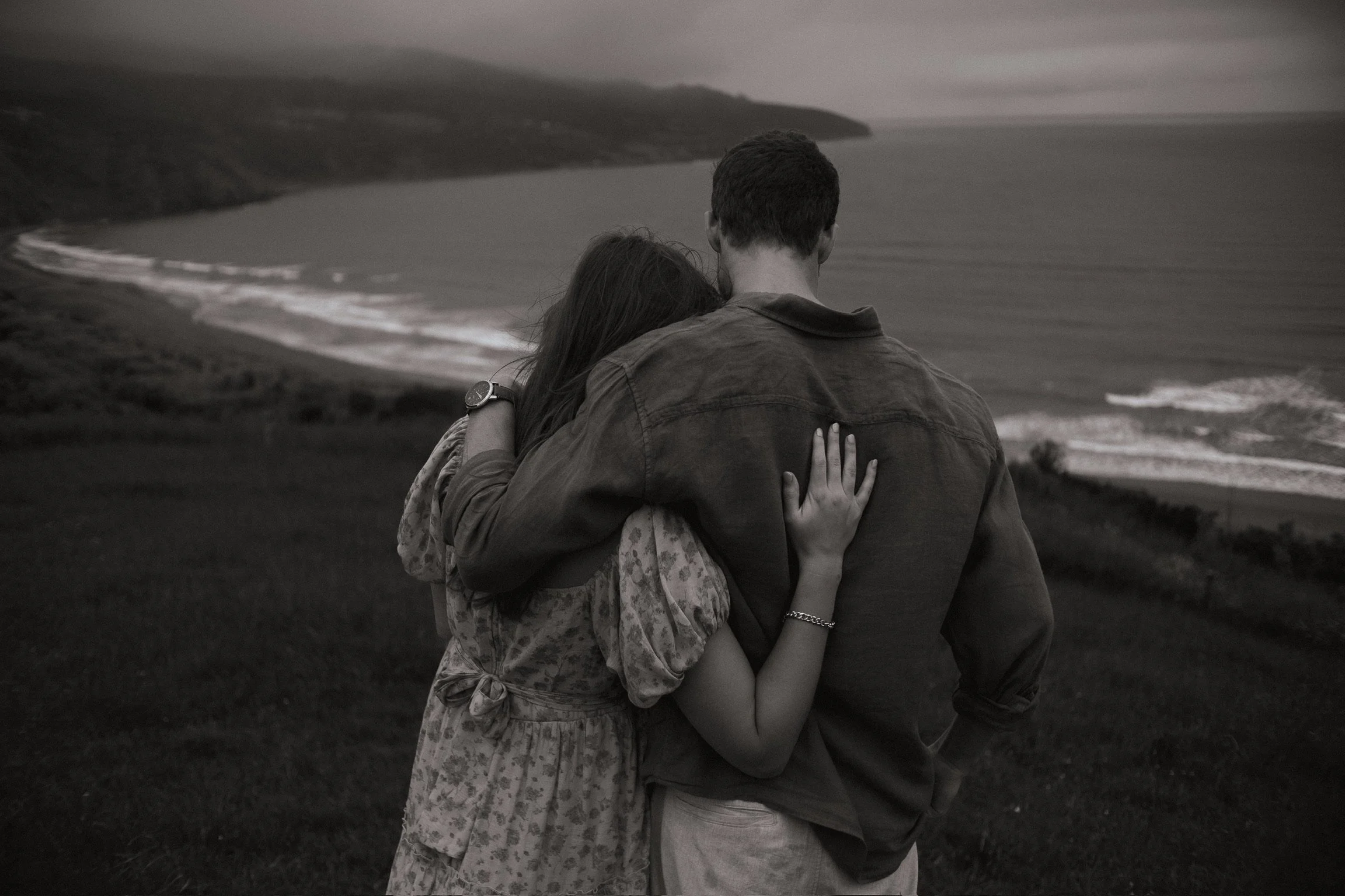 A couple standing close together on a hill overlooking the ocean, with the woman's head resting on the man's neck, in a black and white photo. Couples photography Raglan New Zealand. Engagement photography Hamilton New Zealand. 