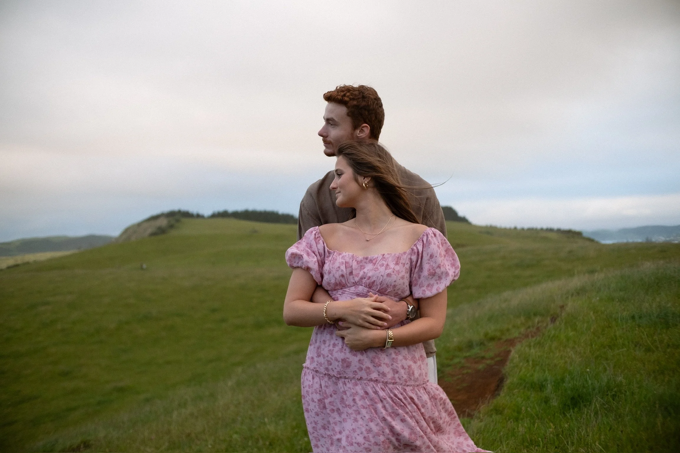 A couple stands in a green field on a cloudy day, with the man embracing the woman from behind. The woman is wearing a pink patterned dress with puffed sleeves, and the man is dressed in a brown shirt. They appear relaxed and content, with hills and 