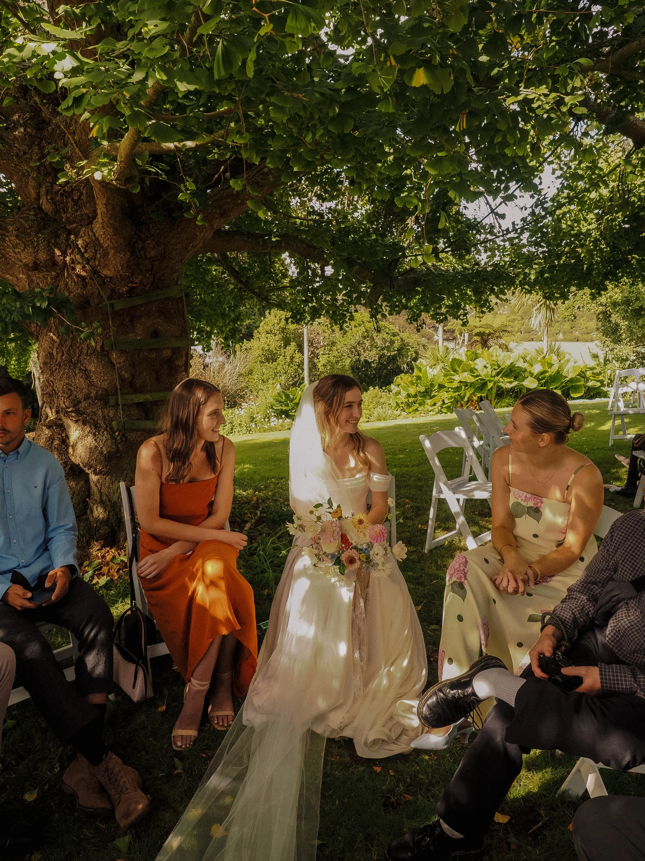 A bride in a white gown with a veil sitting outdoors under a large tree, surrounded by friends, during a wedding ceremony. The bride is holding a bouquet of flowers and smiling at a woman in a cream-colored dress with floral patterns.