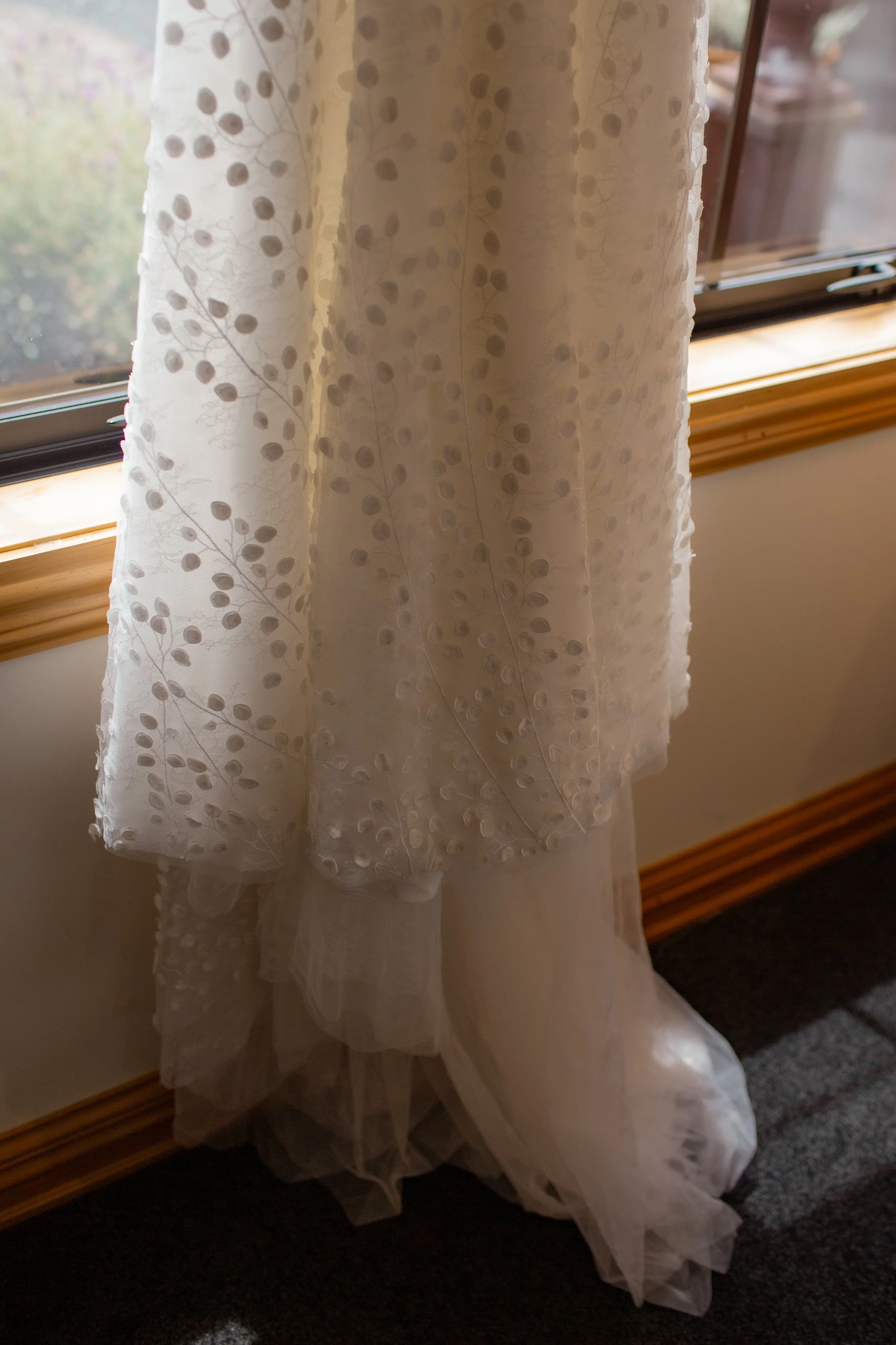 A white wedding dress with floral embroidery hanging by a window.  Wedding Photography Waikato New Zealand. Wedding Photography Hamilton New Zealand. Northland Wedding Photography