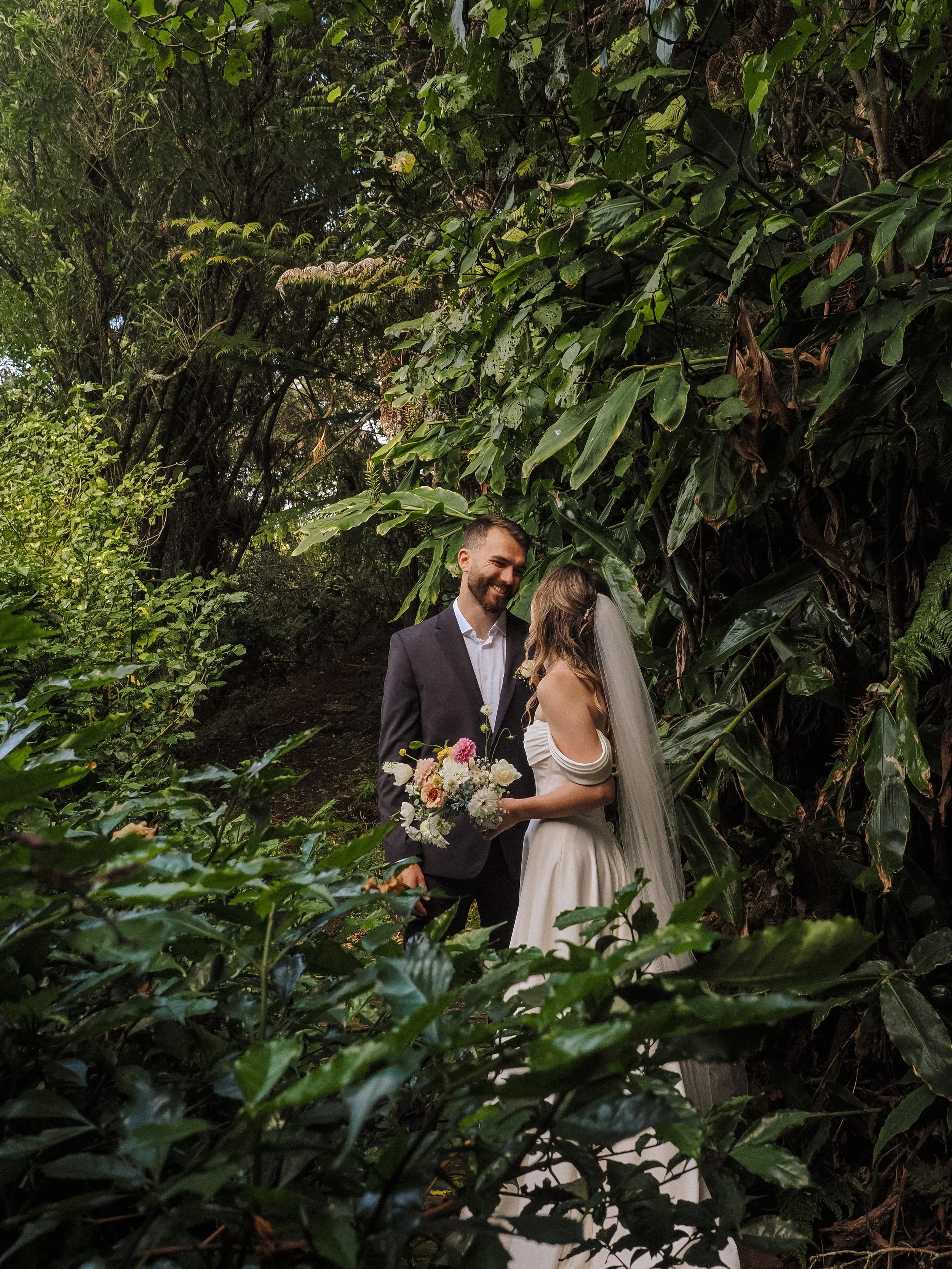 A newlywed couple is standing close together in a lush, green outdoor setting, sharing a joyful moment. The bride is wearing a white off-the-shoulder wedding dress with a long veil, holding a bouquet of flowers. The groom is dressed in a dark suit an