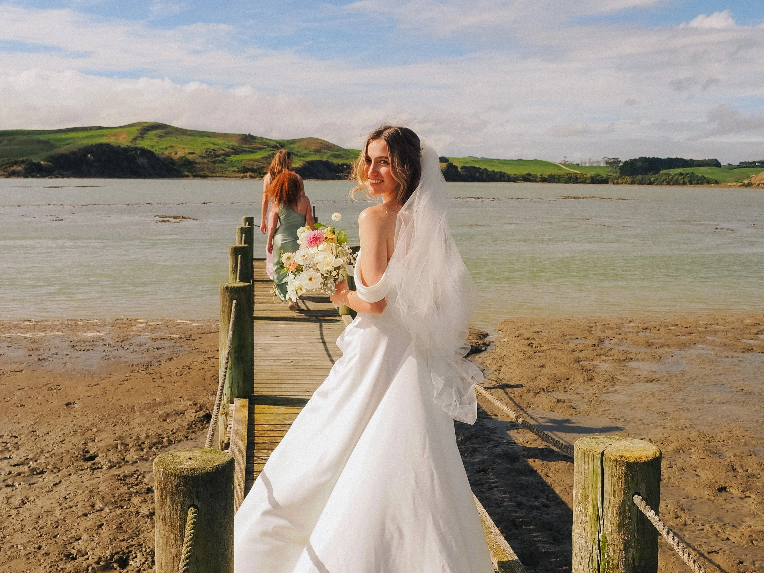 Bride in a white wedding gown holding a bouquet of flowers on a wooden pier by the water, with friends walking behind her on a scenic beach.  Wedding Photography Waikato New Zealand. Wedding Photography Hamilton New Zealand. Northland Wedding Photo