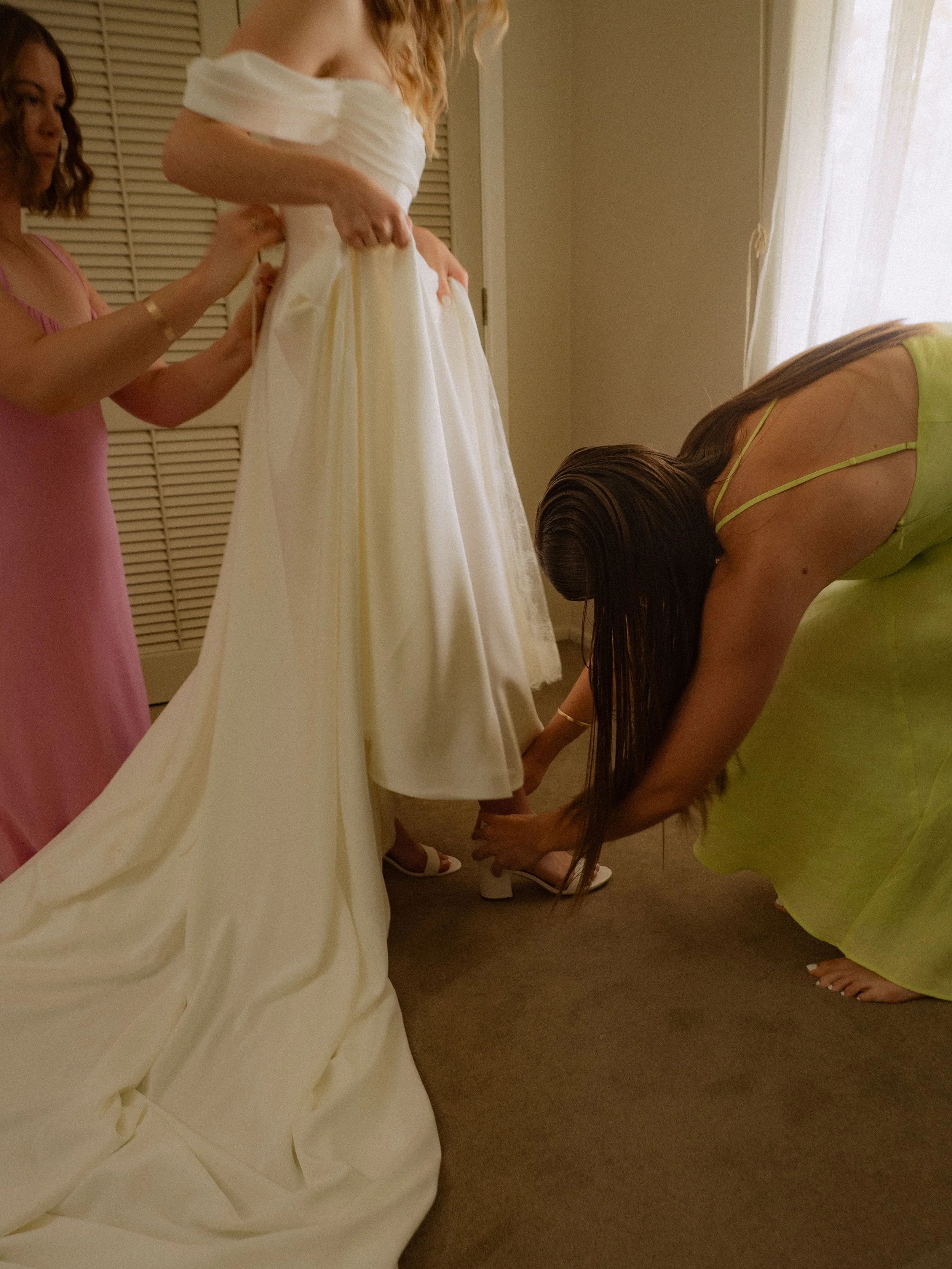 Women helping a bride put on her wedding shoes in a room with soft lighting.  Wedding Photography Waikato New Zealand. Wedding Photography Hamilton New Zealand. Northland Wedding Photography